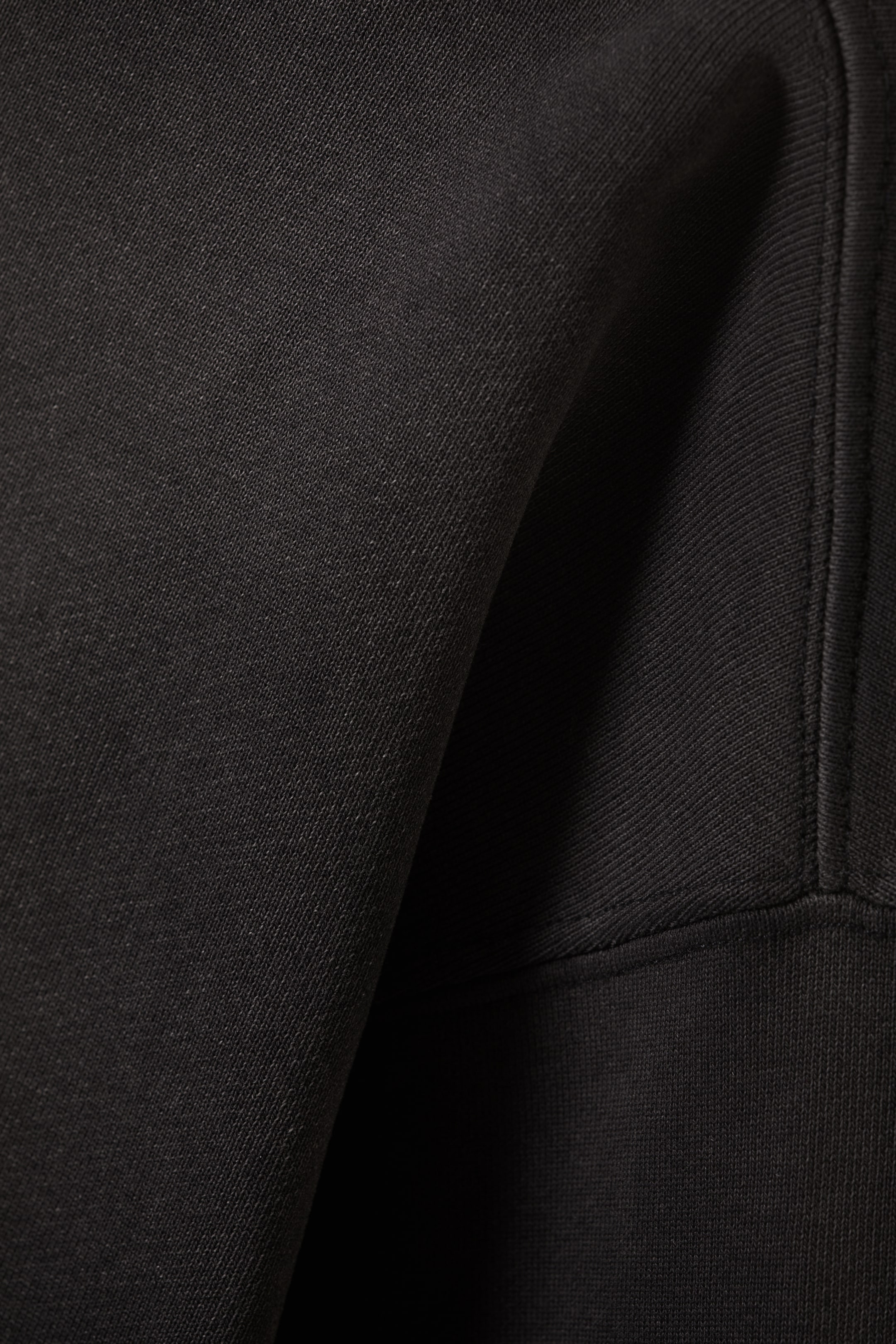 Visualizza immagine più grande: Una sezione di tessuto a maglia nera viene mostrata in primo piano, rivelando la sua sottile texture diagonale e le morbide pieghe. Una cucitura verticale prominente corre lungo il lato destro, collegandosi a una fascia a coste orizzontali che forma un polsino o un orlo.