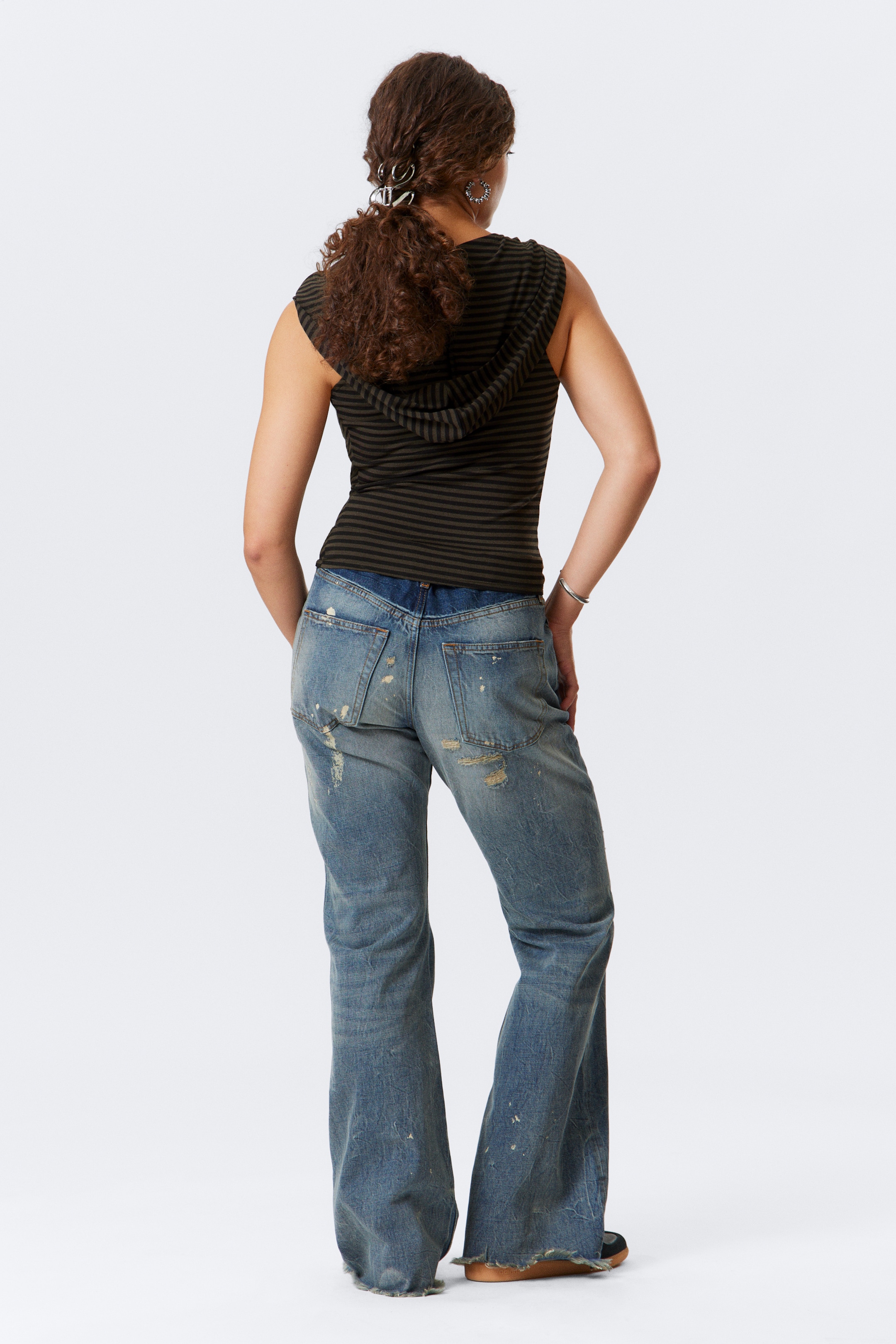 Ingrandisci l'immagine: Un top con cappuccio senza maniche, visto da dietro, presenta strisce orizzontali verde oliva scuro e nere. Viene indossato con jeans in denim azzurro chiaro con effetto consumato che mostrano un retro a vita bassa e una gamba ampia e svasata con un orlo grezzo.