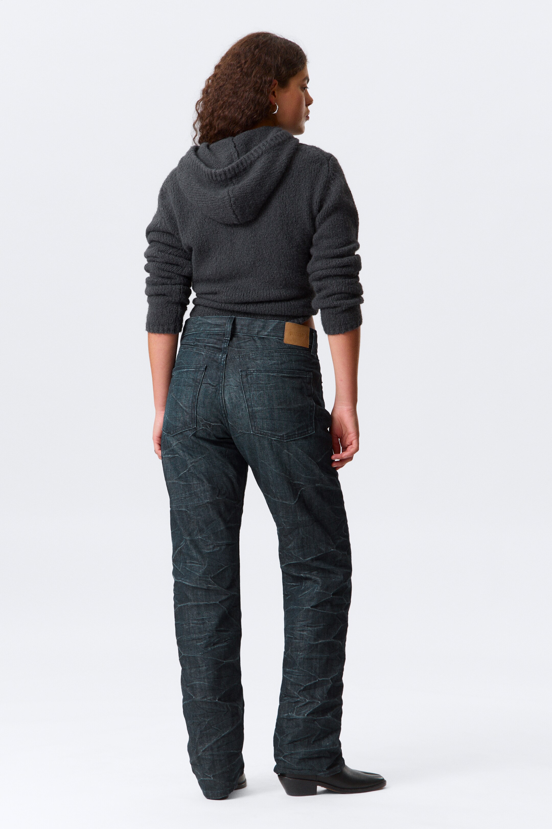 Se større billede: En mørkegrå, struktureret striktrøje med hætte og mørkvaskede jeans ses bagfra. Den afkortede hættetrøje har en hætte og lange ærmer, mens de lige jeans fremviser en krøllet tekstur, baglommer og et lysebrunt mærke på linningen.