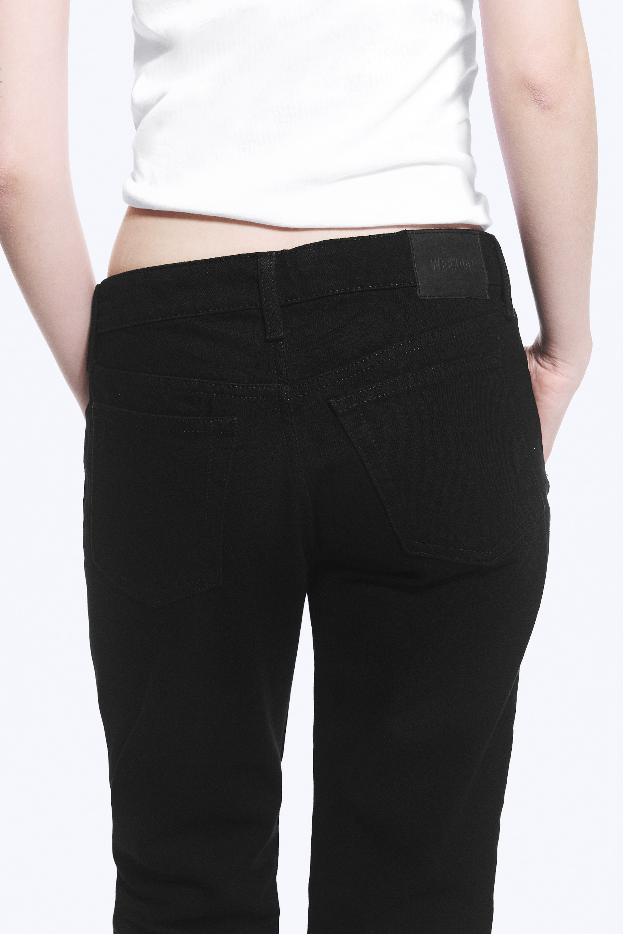 Agrandir l'image: Le jean noir est présenté de dos, avec une ceinture dotée de passants pour ceinture et une étiquette de marque noire. Deux poches arrière plaquées sont visibles avec des coutures ton sur ton, et les mains reposent dans les poches latérales, mettant en valeur la coupe ajustée autour de la taille et des hanches.
