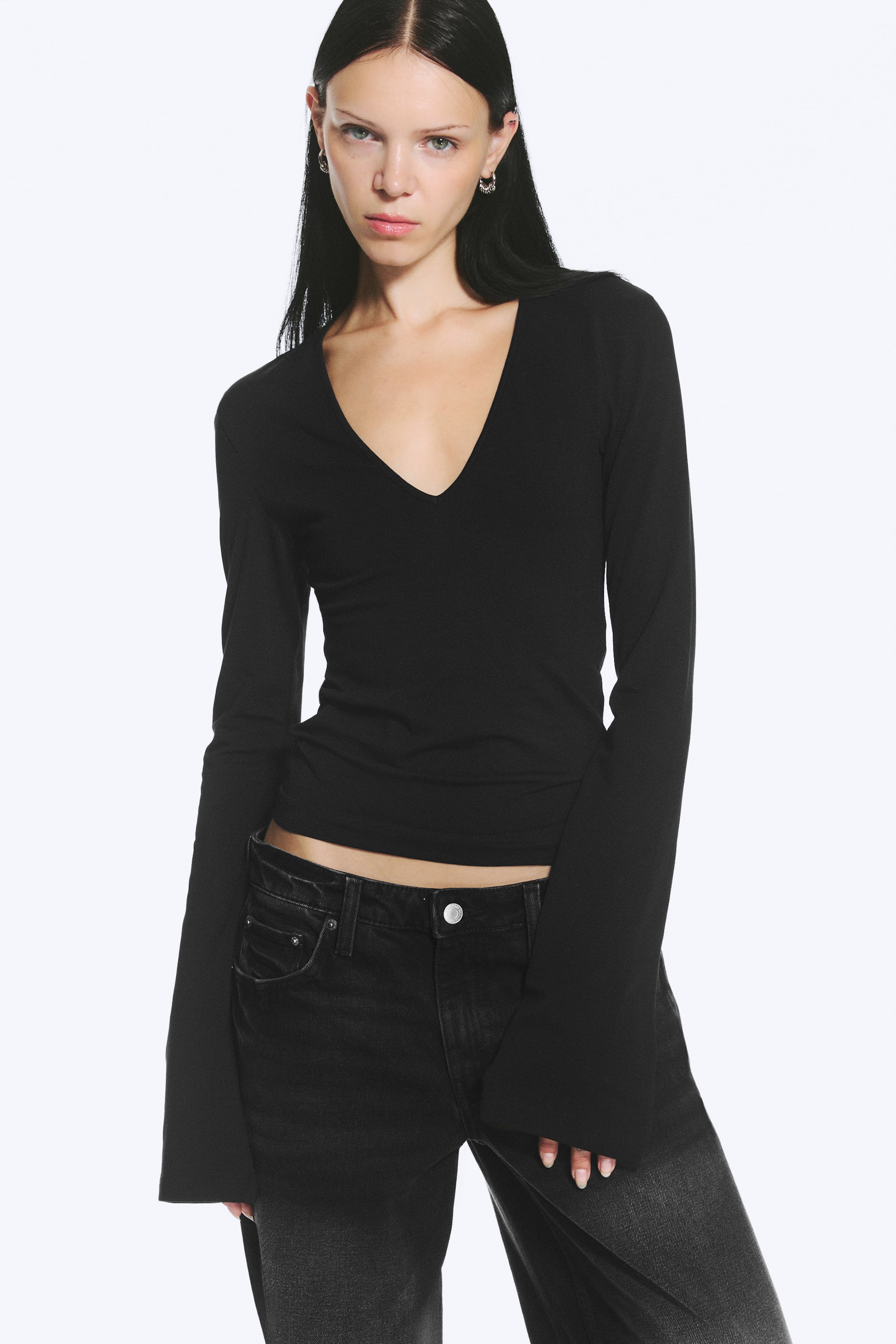 View larger image: A black, slim-fit, long-sleeved top featuring a deep V-neckline and extra-long sleeves is worn by the model. The top is styled with a pair of low-rise black jeans.