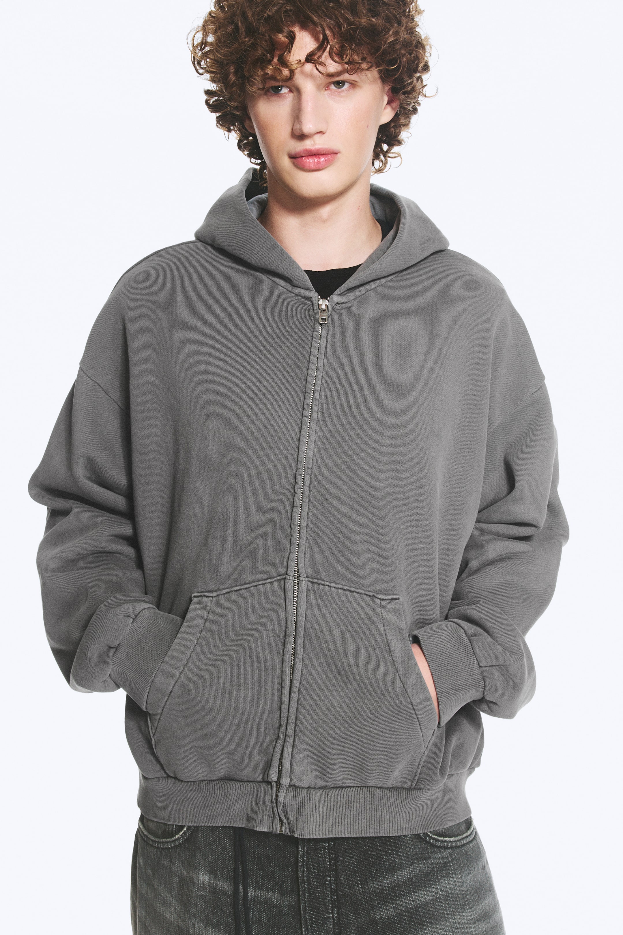 Ver imagen más grande: Una sudadera con capucha de cremallera completa en gris carbón lavado presenta un corte relajado y oversize en una vista frontal. Cuenta con una cremallera metálica prominente, bolsillos canguro divididos y puños y dobladillo acanalados, con una camiseta negra visible en el escote.