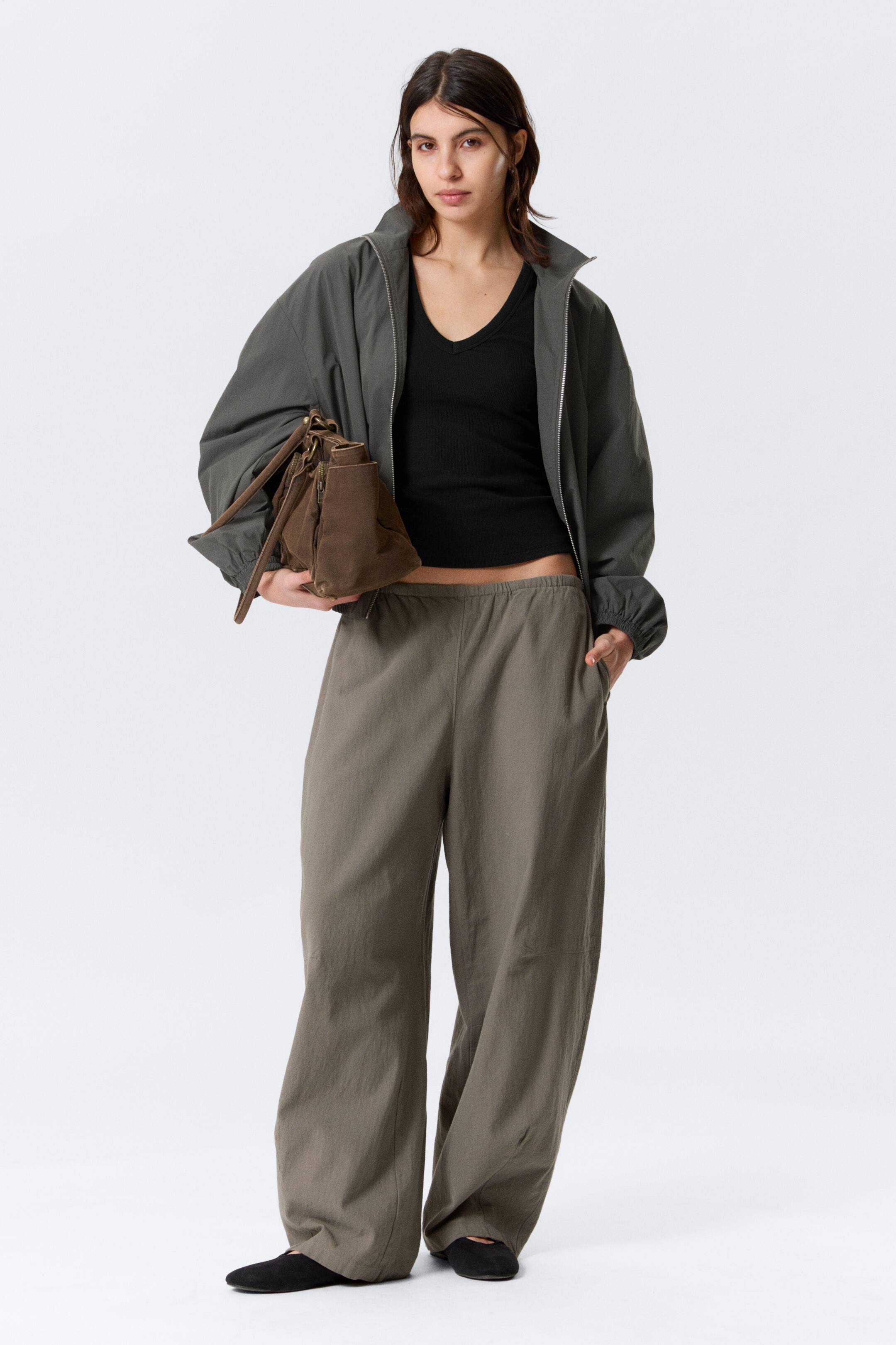 View larger image: Olive green low-rise pull-on trousers, featuring a loose fit, elasticated waistband, and subtle shaping darts at the knees and hem, are being worn by the model, whose hand is resting in a side seam pocket.
