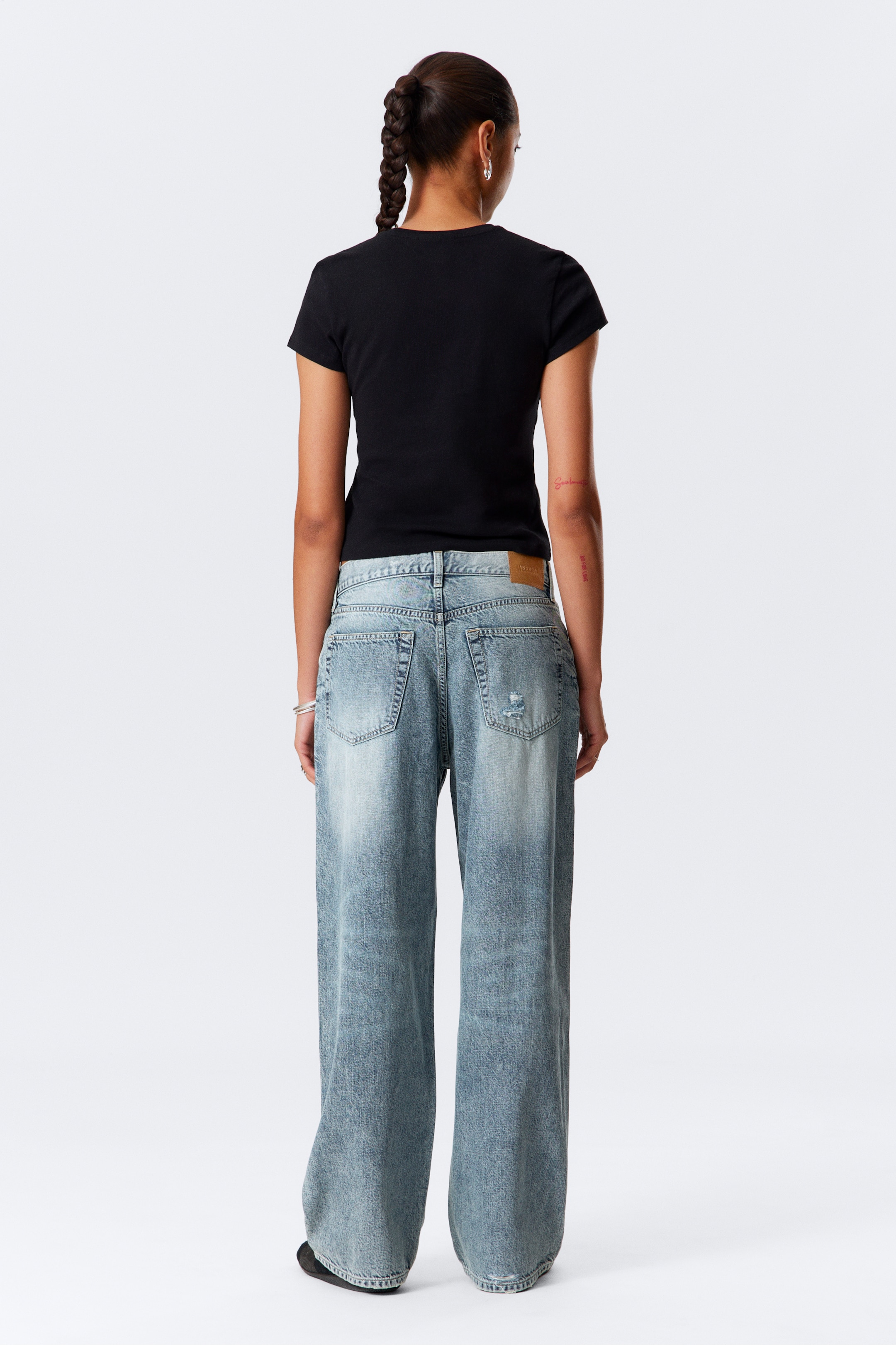 Grösseres Bild ansehen: Ein eng anliegendes schwarzes Kurzarm-Top wird von hinten gezeigt, getragen mit einer hell gewaschenen, locker sitzenden Jeans. Die Jeans verfügt über zwei sichtbare aufgesetzte Gesäßtaschen, einen kleinen Bundaufnäher und abgenutzte Details an der rechten Gesäßtasche, mit weiten Beinen.