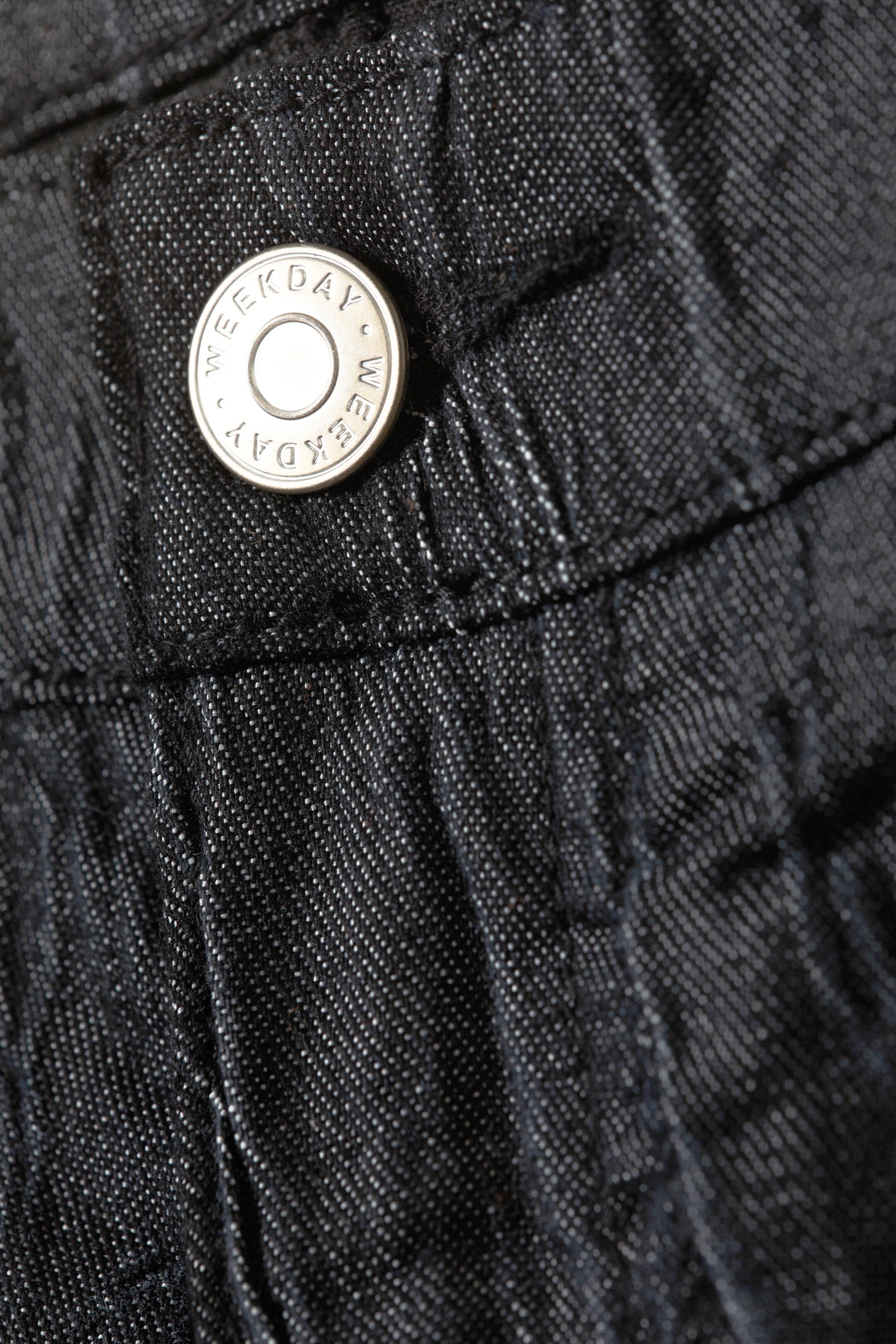 Grotere afbeelding bekijken: De voorkant van een donker denim kledingstuk heeft een zilverkleurige knoop met 'WEEKDAY' rondom de rand gegraveerd. De donkere denim stof vertoont een zichtbare weefstructuur en subtiele lichte spikkels, met stiksels die de knoop bevestigen en horizontale naden vormen.