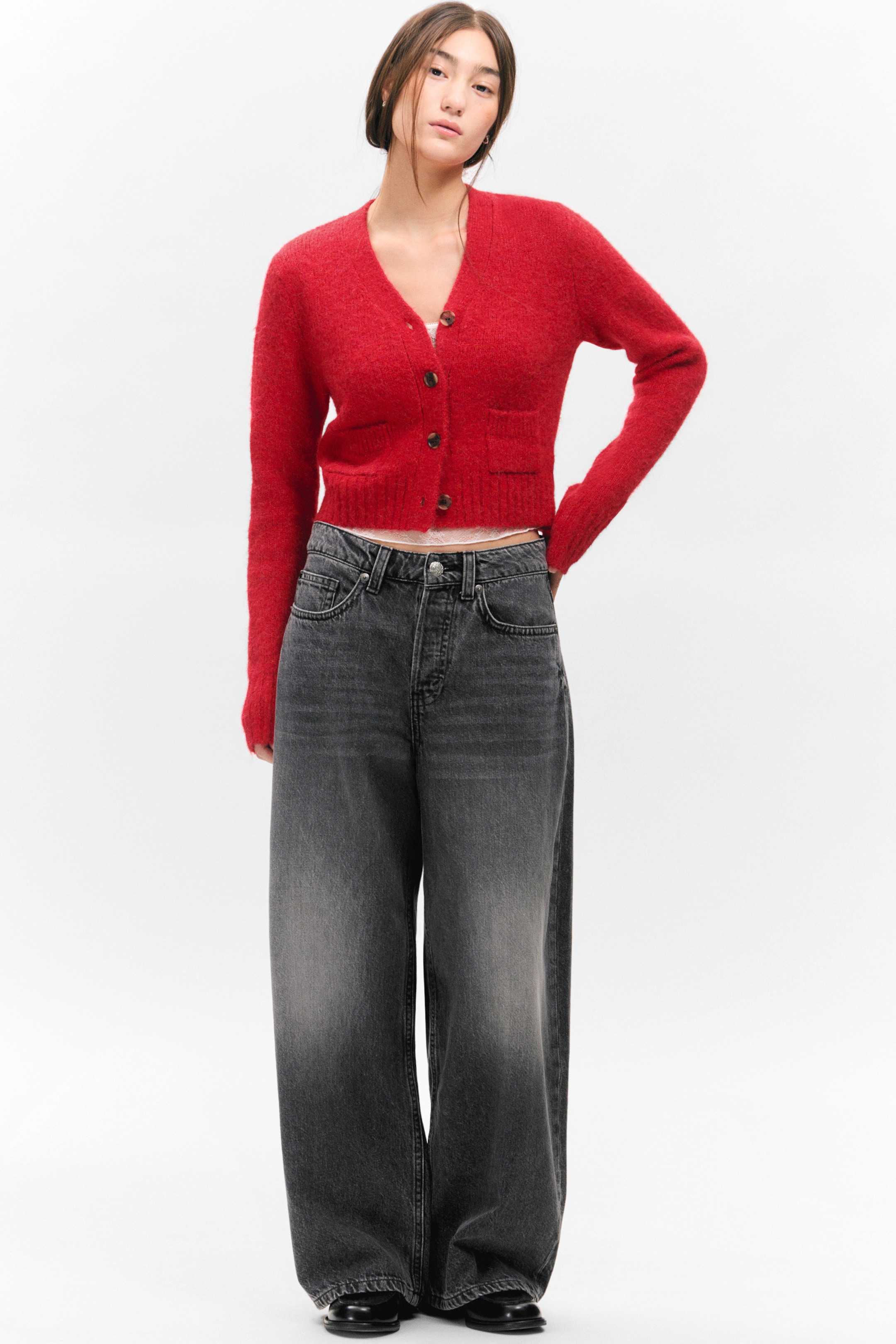 Größeres Bild ansehen: Eine rote V-Ausschnitt-Strickjacke, die mit gerippten Manschetten und einem gerippten Saum, zwei aufgesetzten Vordertaschen und einer Knopfleiste mit braunen Knöpfen ausgestattet ist, wird vom Model getragen. Die Strickjacke ist teilweise zugeknöpft und wird über einem hellen Oberteil getragen.