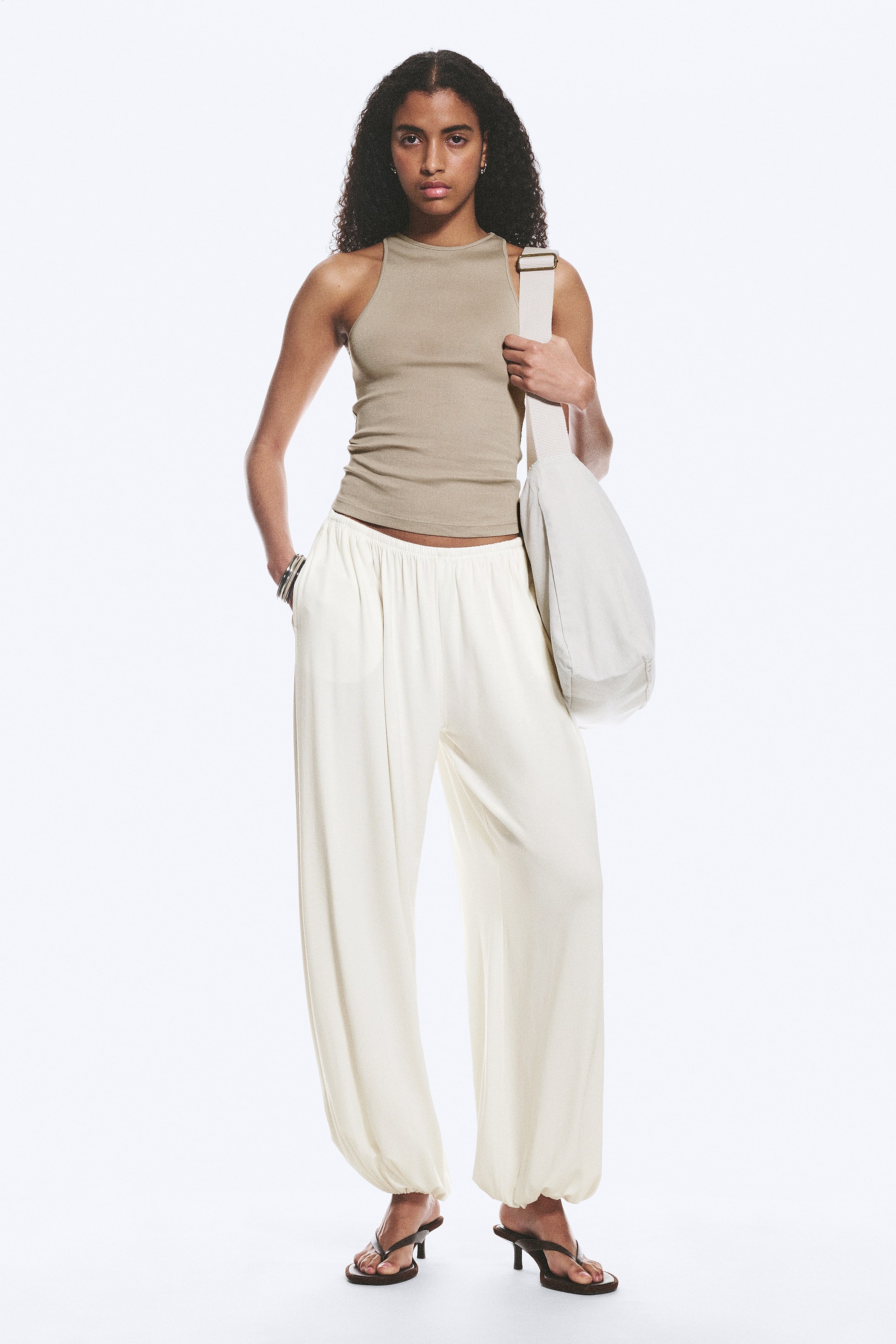 View larger image: Cream loose-fit trousers featuring an elasticated mid-rise waistband, voluminous balloon-shaped legs, and elasticated cuffs are being worn by the model, who is placing one hand into a side seam pocket.