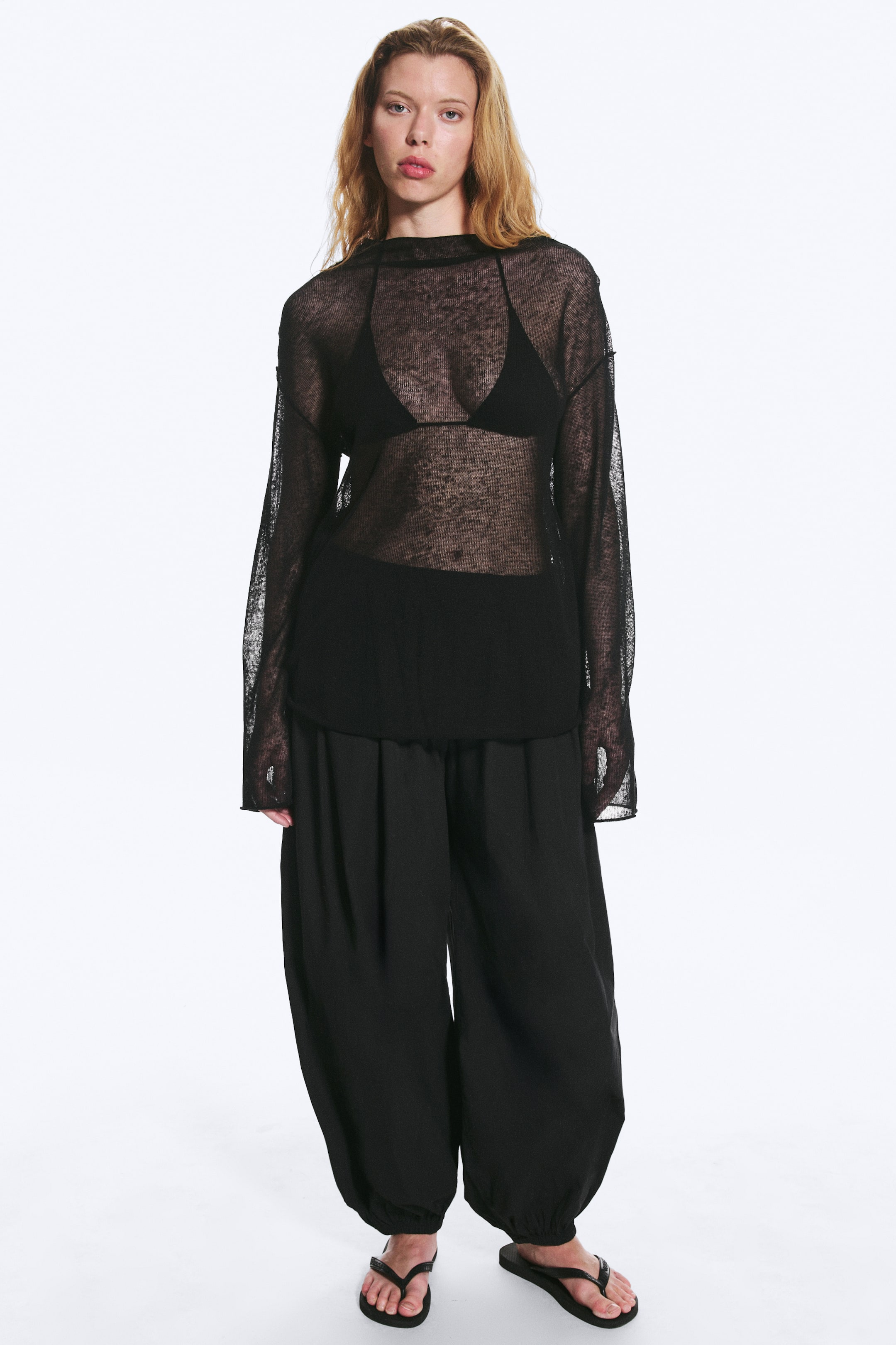 Agrandir l'image: Un haut noir en maille transparente à manches longues est porté, avec un col montant, une coupe ample et des poignets à bords bruts, révélant un soutien-gorge noir en dessous. Un pantalon noir à jambes larges avec chevilles froncées est également porté, offrant une silhouette décontractée et ample.