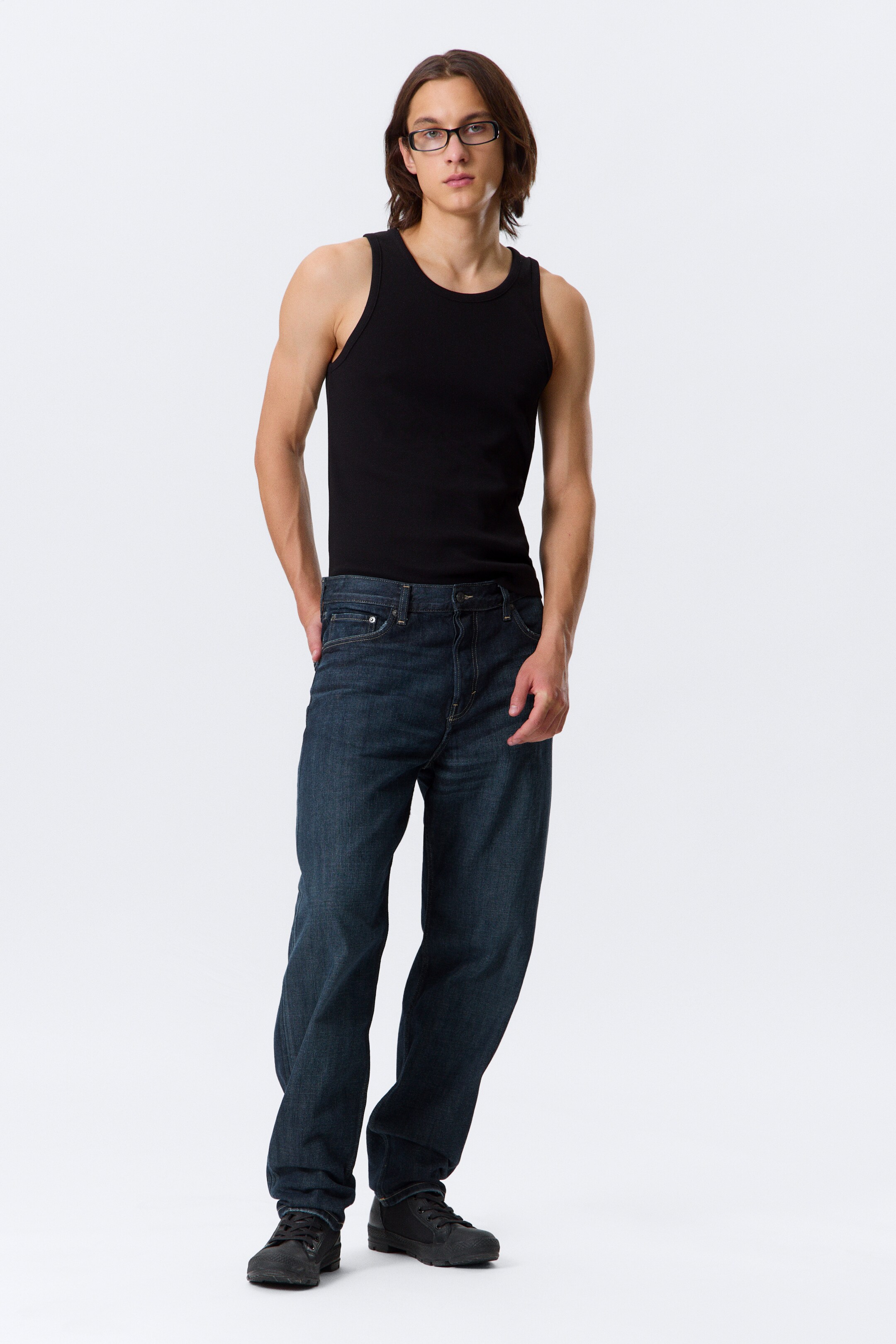 View larger image: A black ribbed tank top is featuring a round neck and wide shoulder straps, fitting snugly. Dark wash, straight-leg jeans are showcasing a relaxed fit with some creasing.