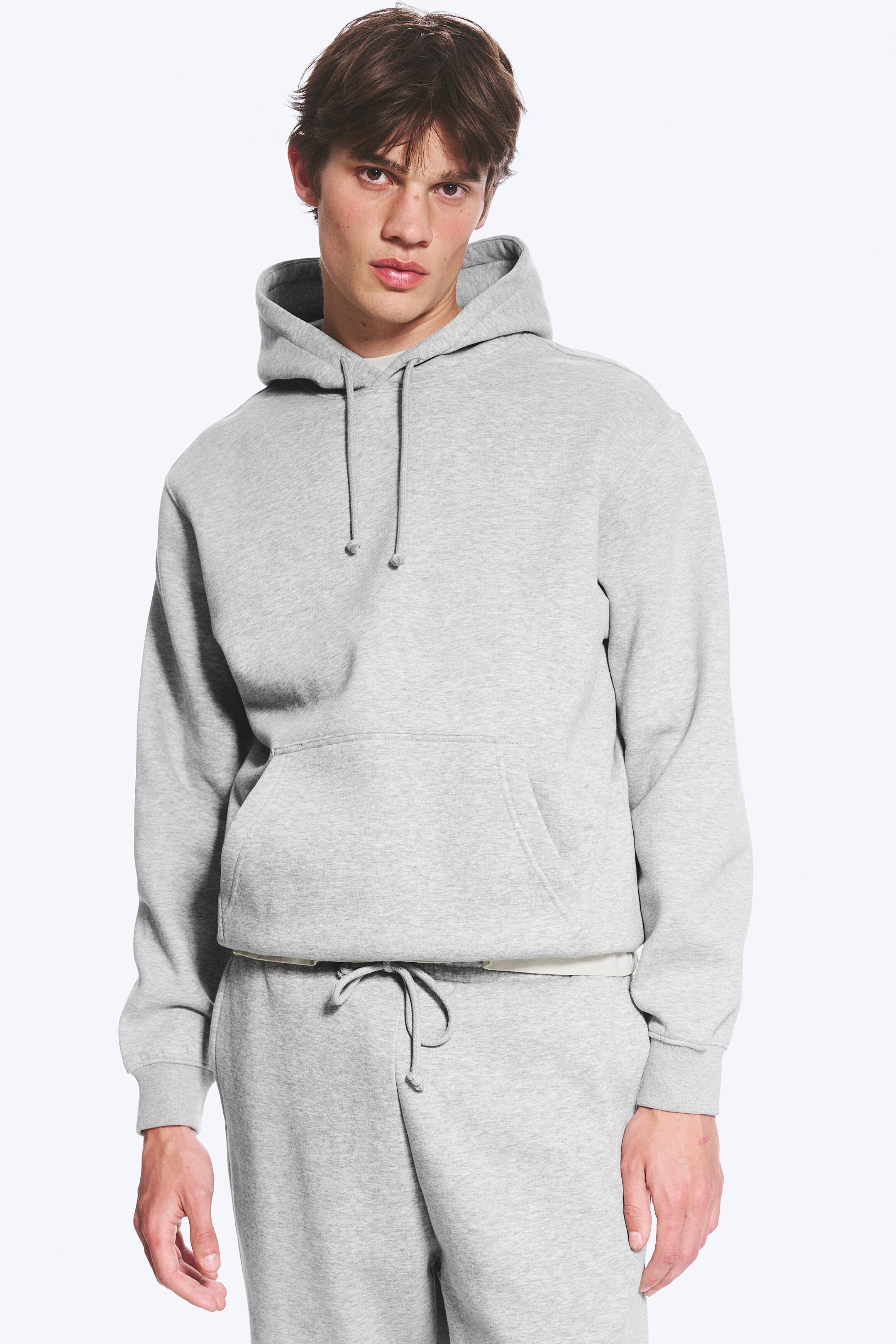Größeres Bild anzeigen: Ein hellgrauer Hoodie wird von vorne präsentiert, ausgestattet mit einer Kapuze mit Kordelzug, einer großen Kängurutasche und gerippten Bündchen und Saum. Er zeigt eine lockere Passform mit einer hellen Schicht, die am unteren Saum sichtbar ist.