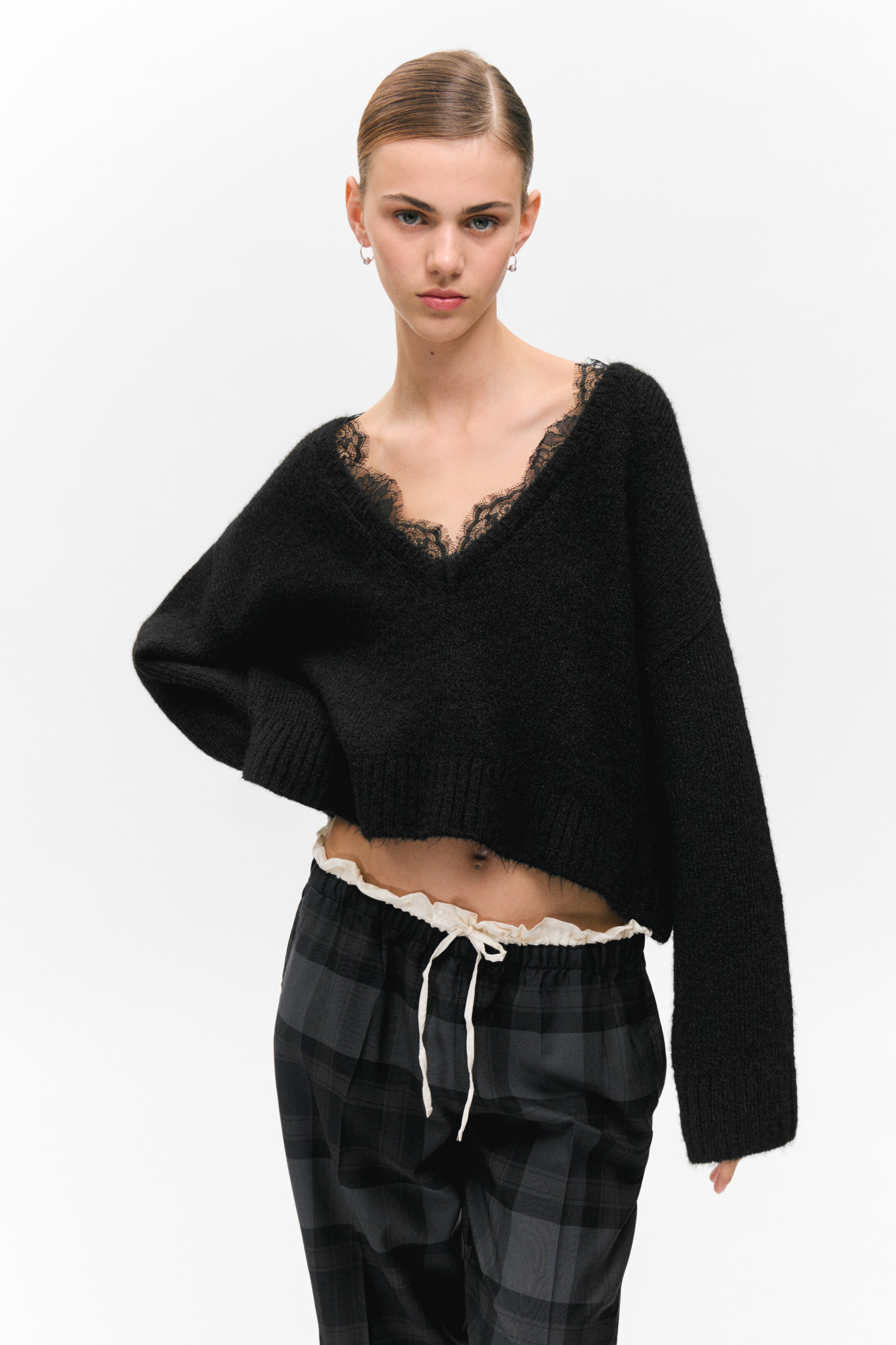 Visualizza immagine più grande: Il maglione nero a V presenta una texture a maglia pelosa e un bordo in pizzo nero lungo lo scollo. È caratterizzato da una lunghezza corta, con un orlo a coste, e le sue maniche lunghe e larghe sono rifinite con polsini a coste, offrendo una vestibilità rilassata dal davanti.
