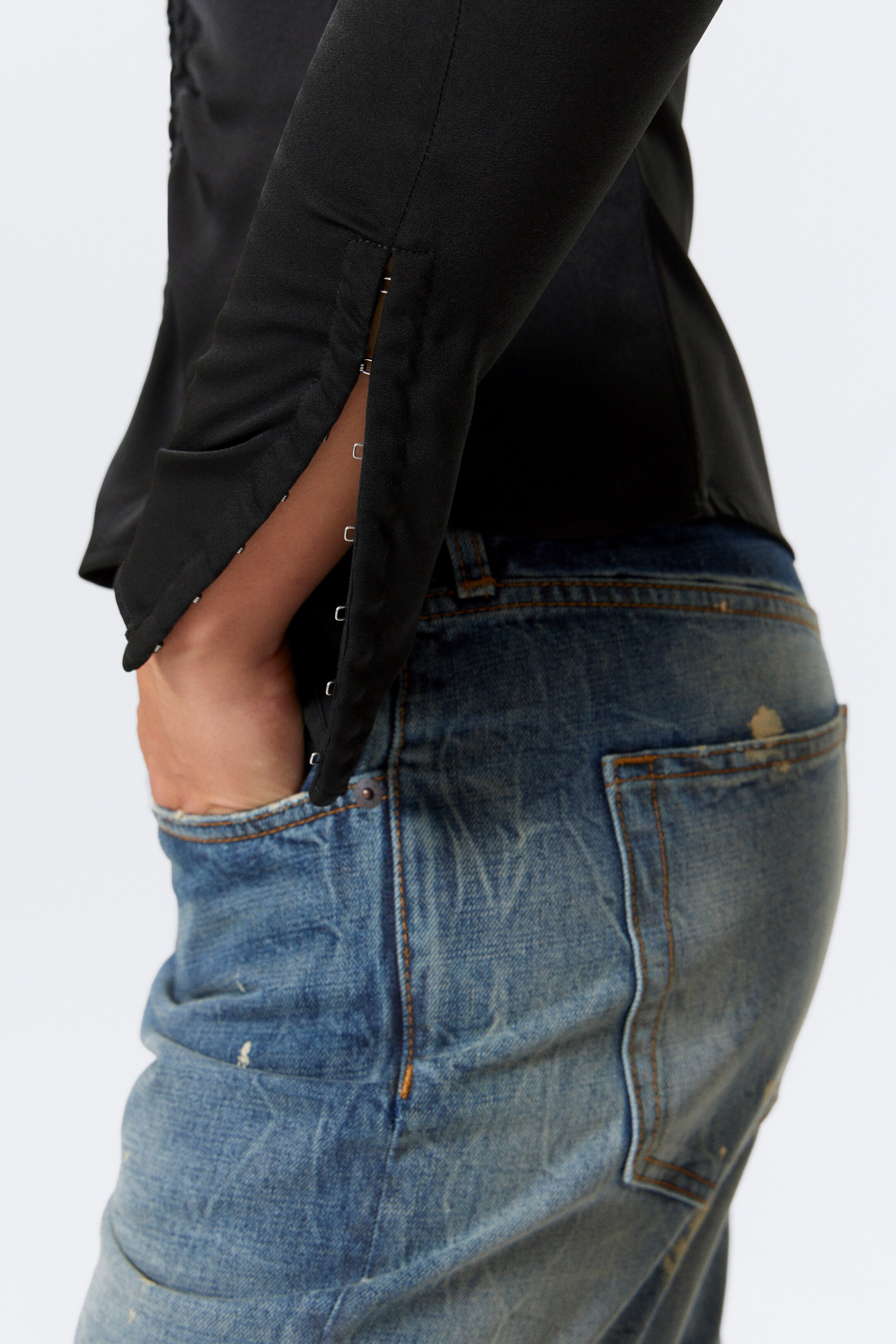 View larger image: A black long-sleeved top is being presented from a side angle, highlighting a split sleeve opening secured with multiple silver-toned hook-and-eye closures. The smooth fabric of the top is being worn with distressed blue jeans, with a hand visible inside a jeans pocket.