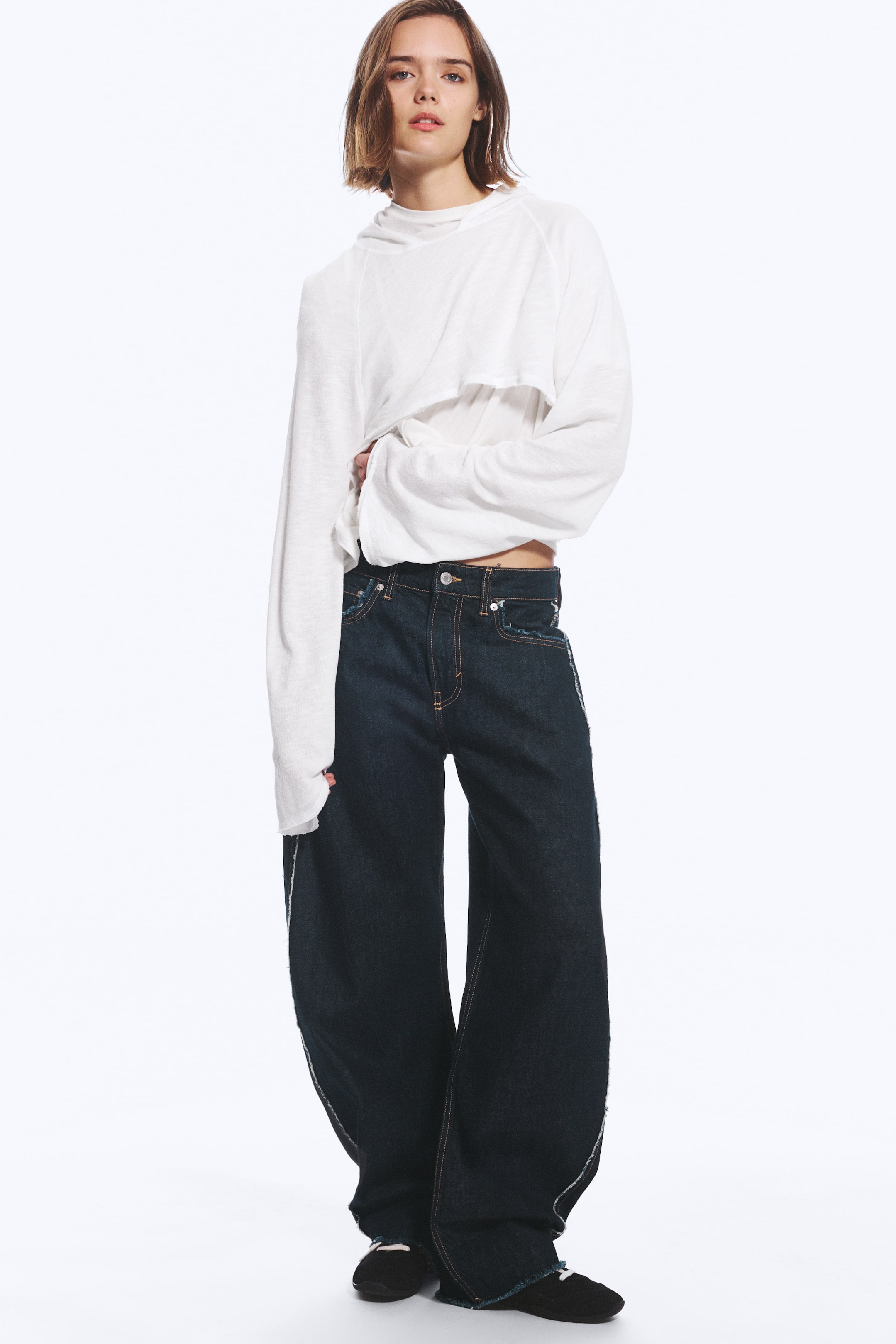 Agrandir l'image: Un jean indigo foncé taille mi-haute, doté d’une silhouette barrel-leg sculptée et de surpiqûres contrastantes à l’extérieur, est porté par le mannequin. Le jean est associé à un top blanc à manches longues superposé.