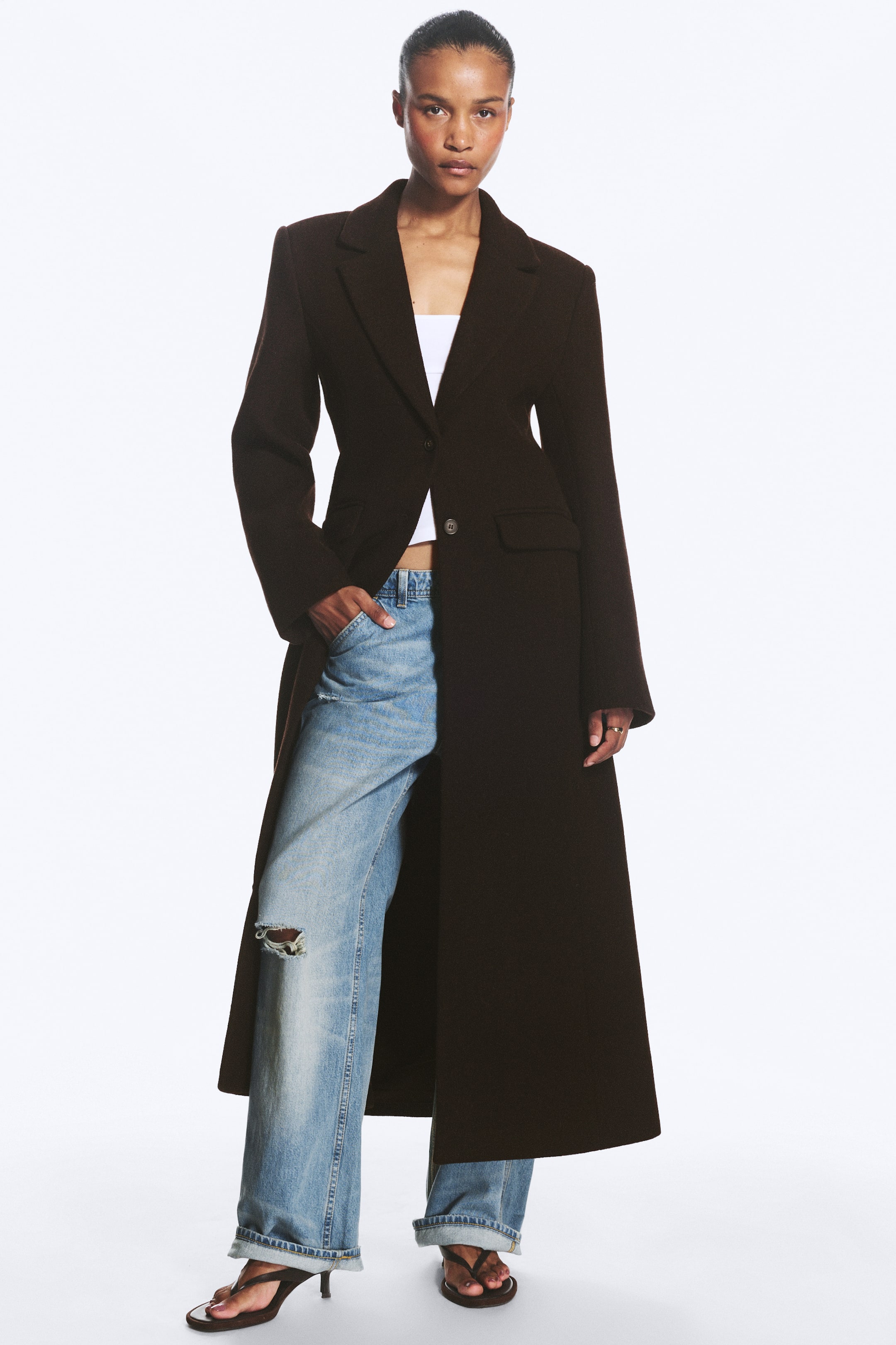 Agrandir l'image: Un manteau marron foncé longueur totale met en valeur une coupe ajustée, des revers crantés, une fermeture croisée simple avec un seul bouton visible et des poches à rabat. Le manteau est porté ouvert, révélant un haut blanc et un jean bleu clair superposé en dessous.