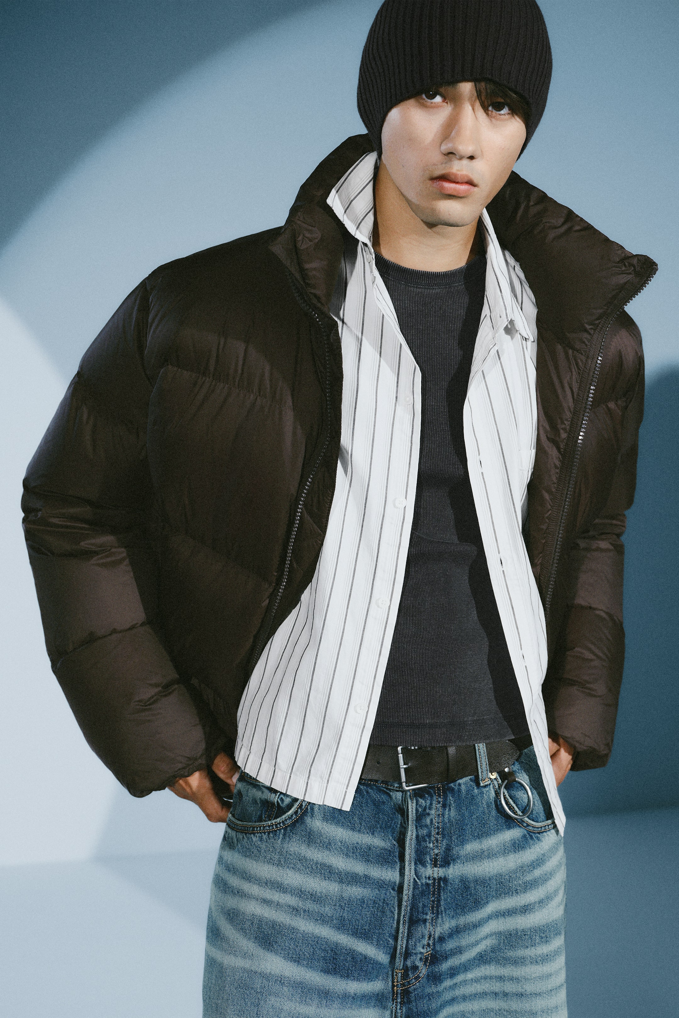 View larger image: A dark brown puffer jacket featuring horizontal quilting and a stand-up collar is worn open by the model. The jacket is layered over an unbuttoned striped shirt, a dark grey T-shirt, and blue jeans.