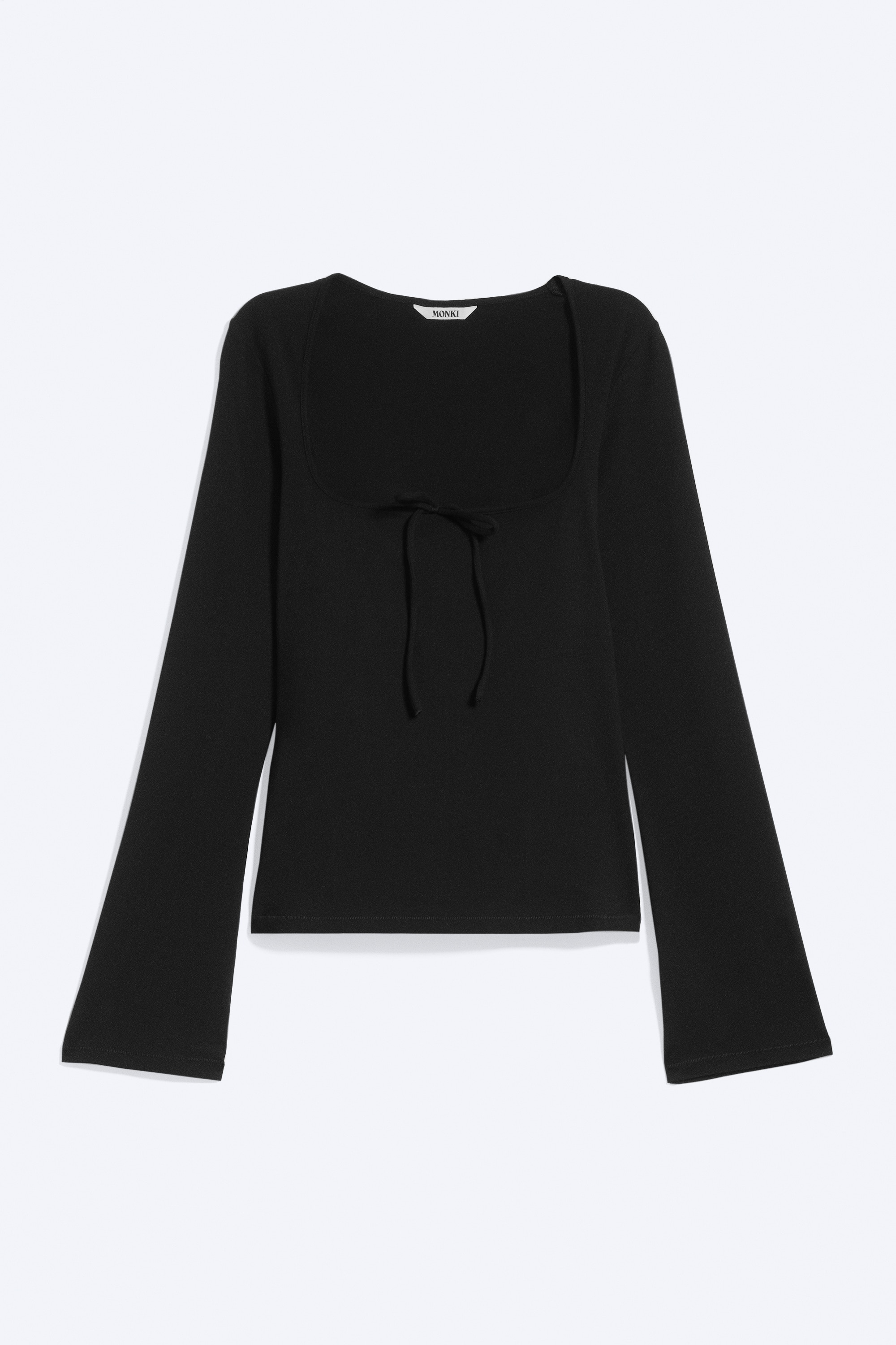 La modelo lleva una blusa negra de manga larga y corte ajustado, con escote cuadrado, detalle de lazada frontal y mangas ligeramente acampanadas. La blusa se combina con una falda negra de encaje a juego.