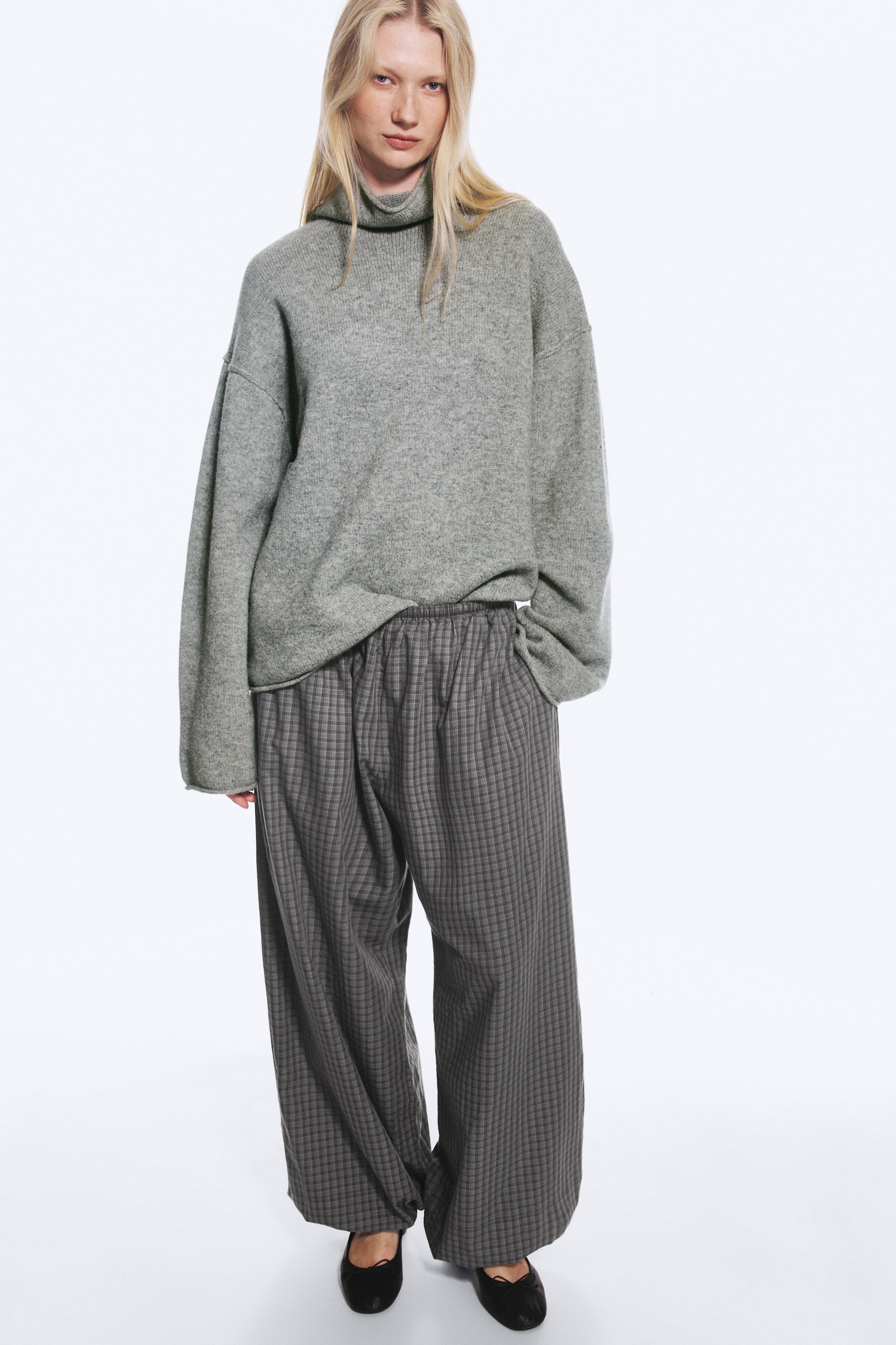 Visualizza immagine più grande: Un paio di pantaloni balloon grigi fantasia, dalla silhouette ampia e voluminosa con polsini elasticizzati, indossati dalla modella. I pantaloni sono abbinati a un maglione a collo alto grigio oversize e scarpe nere basse, con la modella che tiene una mano nella tasca laterale.