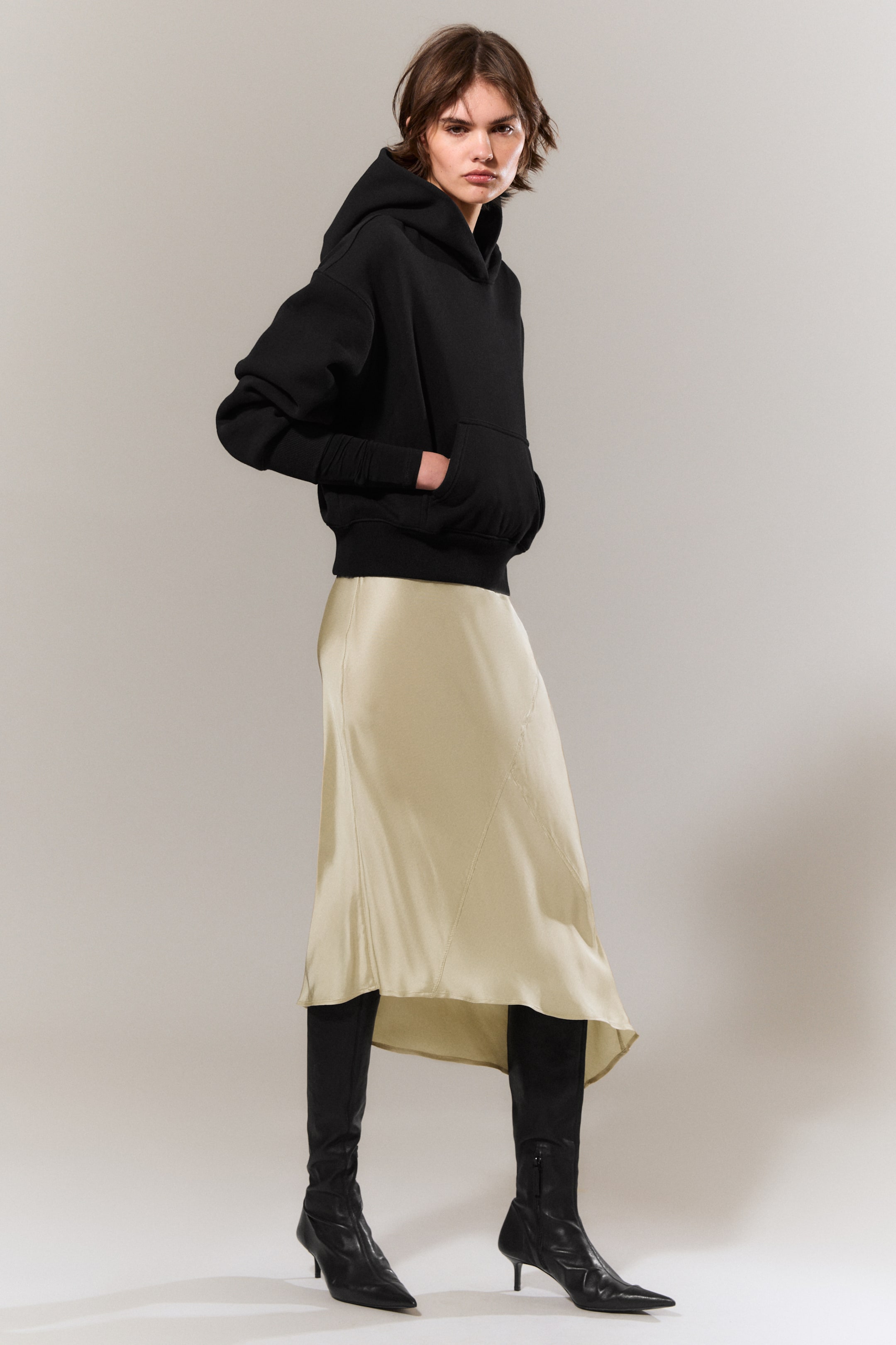 Größeres Bild ansehen: Ein schwarzer Hoodie mit einer kastigen, entspannten Silhouette wird vom Model getragen. Der Hoodie präsentiert überschnittene Schultern, eine geräumige Kapuze, eine Kängurutasche, in der das Model ihre Hände platziert, sowie gerippte Abschlüsse an den Bündchen und am Saum. Er wird über einem hellen Midirock und schwarzen Stiefeln getragen.