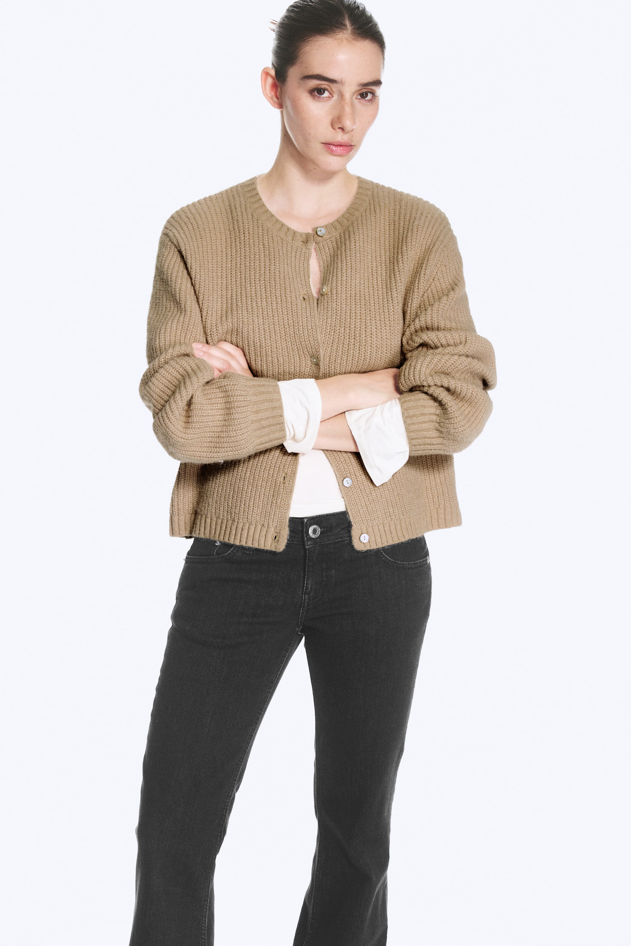 Größeres Bild ansehen: Ein beigefarbener Strick-Cardigan mit Rippstruktur wird vom Model mit verschränkten Armen getragen und über einem weißen Langarmshirt sowie zu dunkelgrauen Jeans kombiniert. Der Cardigan verfügt über einen Rundhalsausschnitt, überschnittene Schultern, eine durchgehende Knopfleiste sowie gerippte Abschlüsse an Ausschnitt, Ärmelbündchen und Saum.
