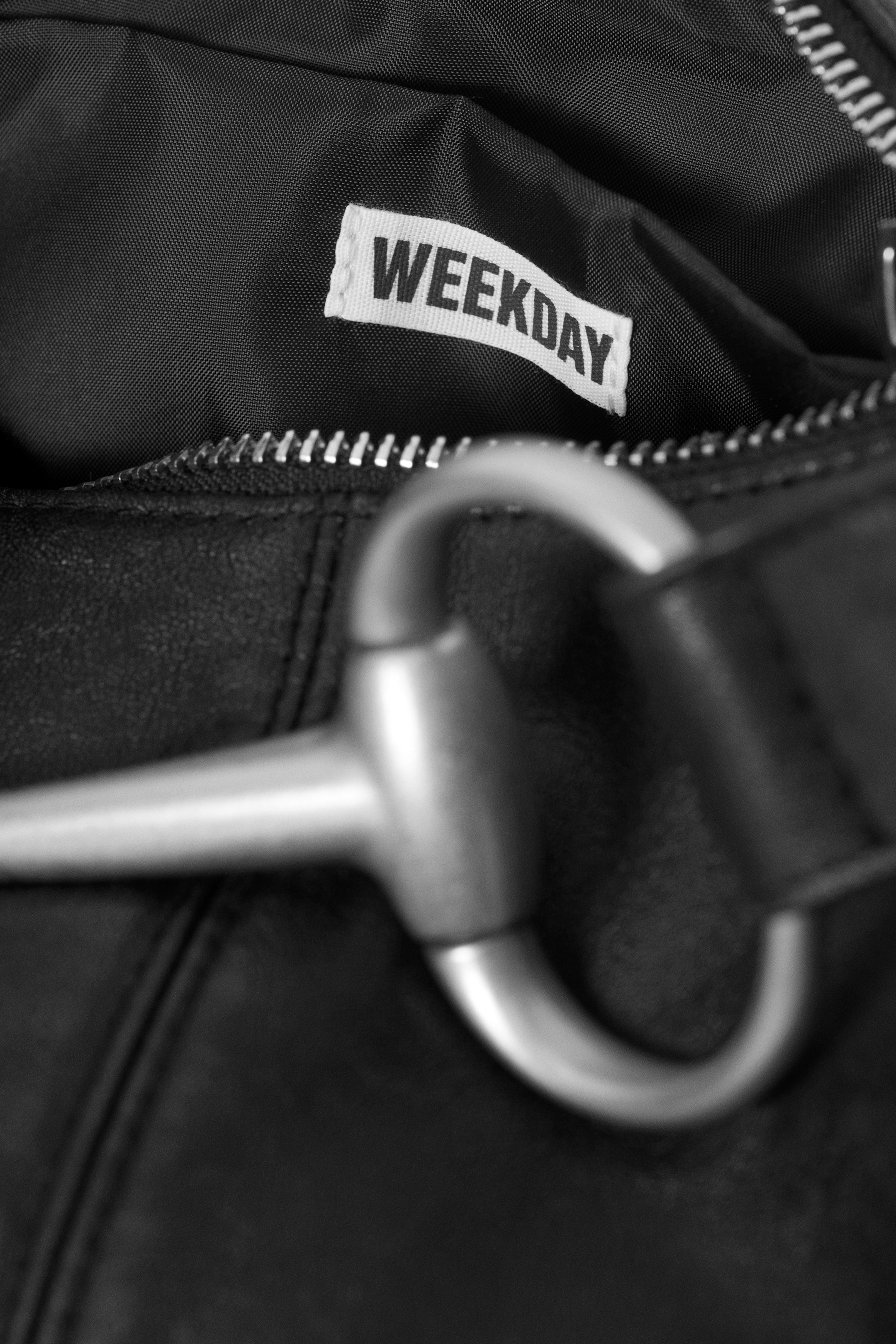 Visa större bild: En svart väska har en vit "WEEKDAY"-etikett insydd i dess svarta tygfoder, synlig genom den delvis öppna silverfärgade metallblixtlåset. Ett stort silverfärgat metallbeslag är också fäst vid väskans texturerade utsida, placerat framträdande i förgrunden.