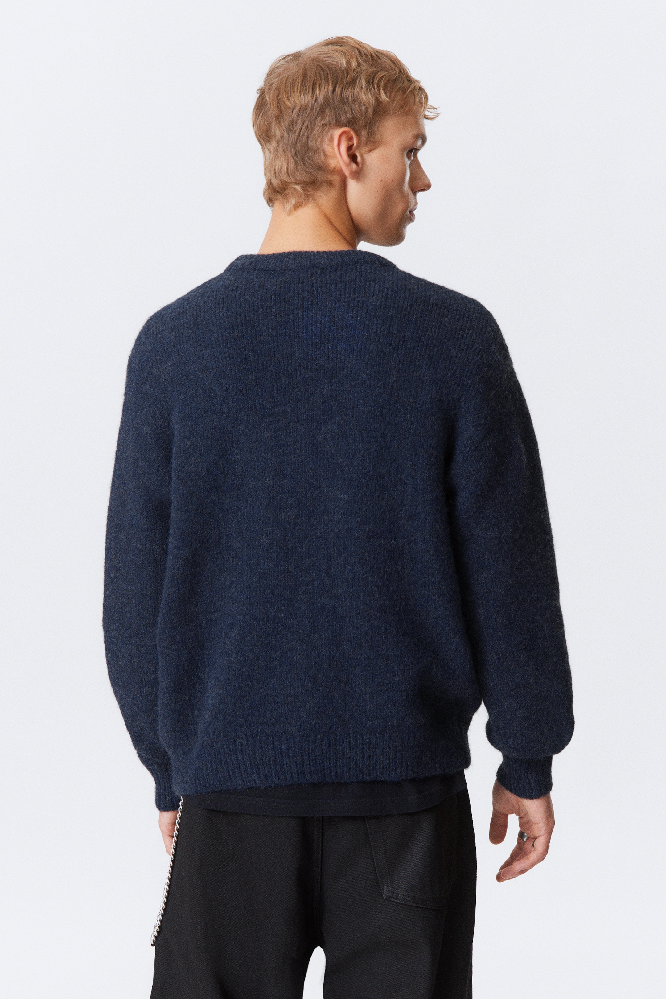 Ver imagen más grande: Un suéter de cuello redondo azul oscuro se está usando, visto desde atrás, ligeramente a la derecha. Presenta un ajuste holgado y una textura peluda, con puños y dobladillo acanalados, y una camiseta negra se está usando debajo, visible en el dobladillo del suéter.