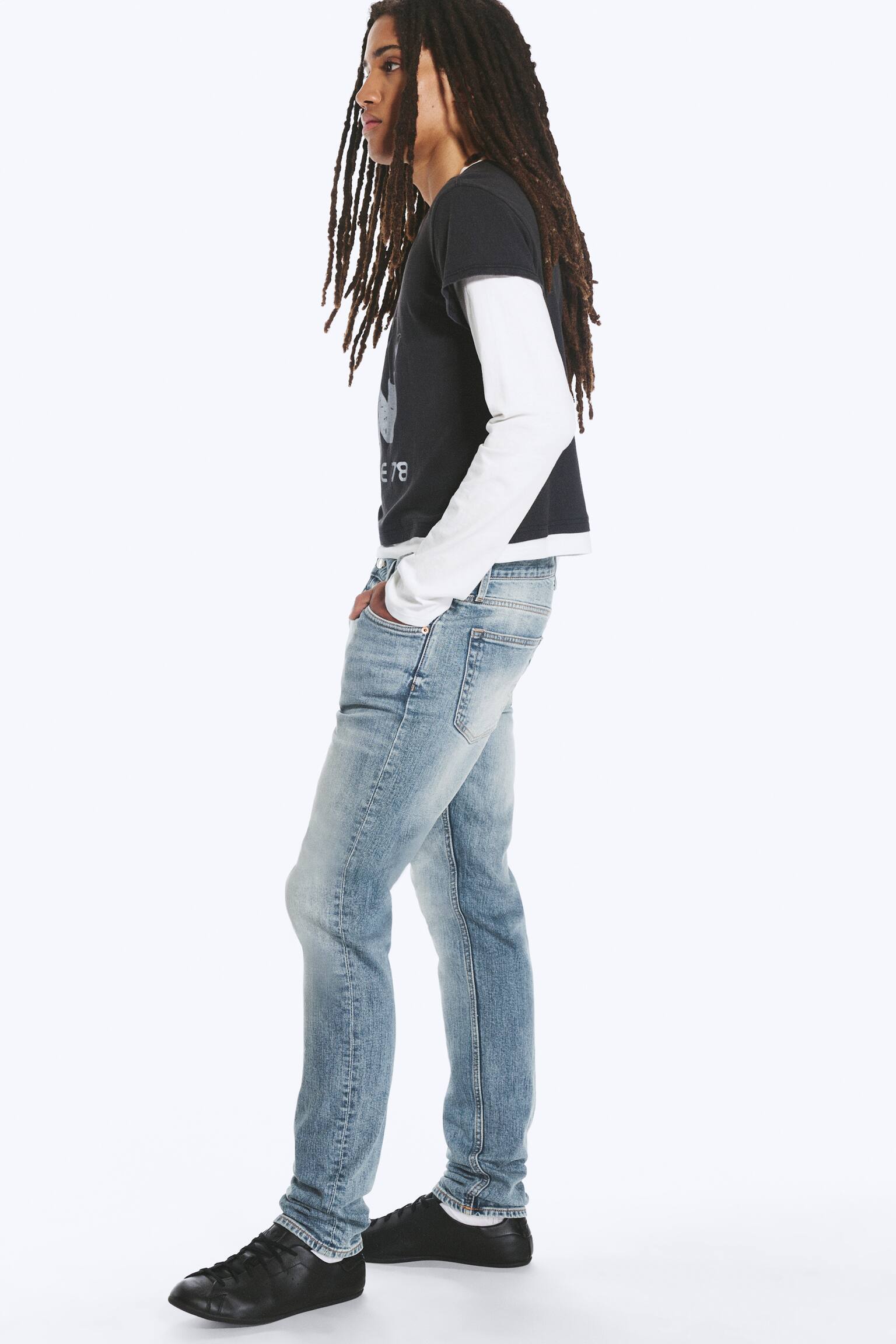 Et par lyse, slim-fit jeans med rette ben bÃ¦res av modellen, som stÃ¥r i sideprofil med Ã©n hÃ¥nd hvilende i frontlommen. Jeansen har lett falming og er stylet med en svart T-skjorte lagt over en hvit langermet skjorte og svarte sneakers. - 1