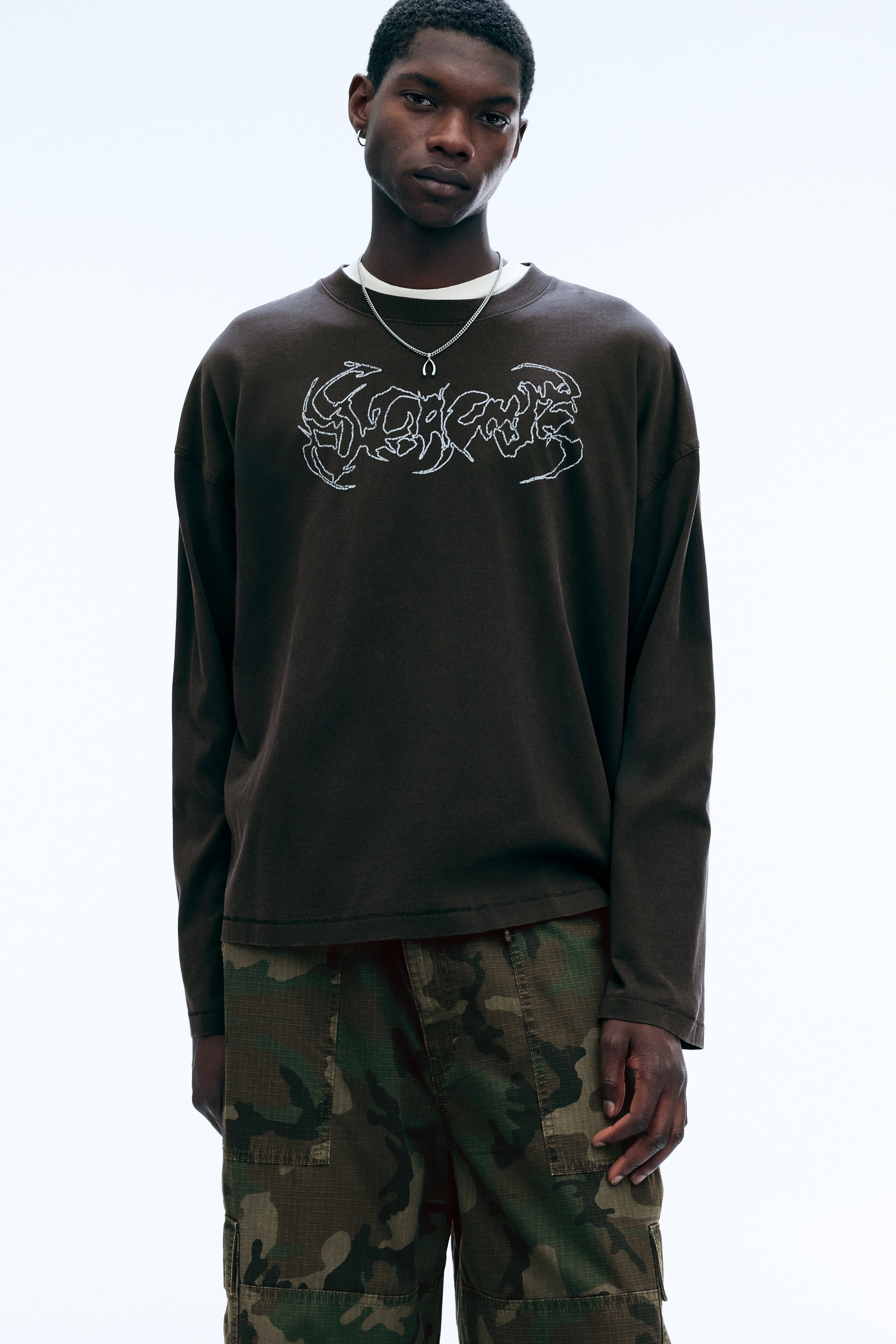 Ce t-shirt oversize Ã  manches longues, de couleur marron foncÃ©, est prÃ©sentÃ© de face, avec un col rond, des Ã©paules tombantes et des manches larges Ã  coupe droite. Un motif graphique abstrait blanc, dans une typographie dentelÃ©e de style mÃ©tal, orne le centre de la poitrine.