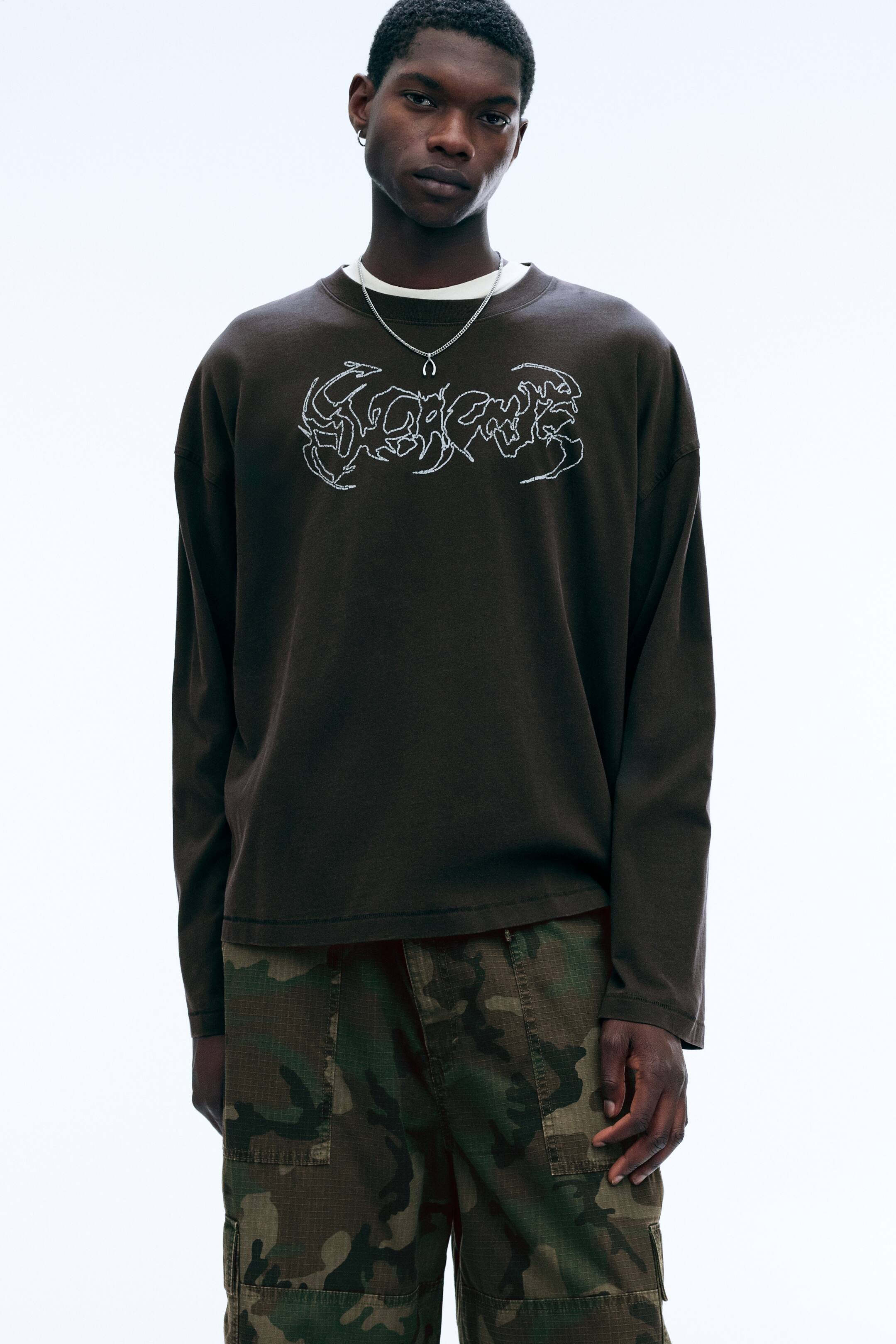 View larger image: An oversized, long-sleeved T-shirt in dark brown is being worn by the model, featuring a crew neck, dropped shoulders, and a white abstract graphic printed on the chest. The product is styled over a white T-shirt and paired with camouflage cargo trousers.