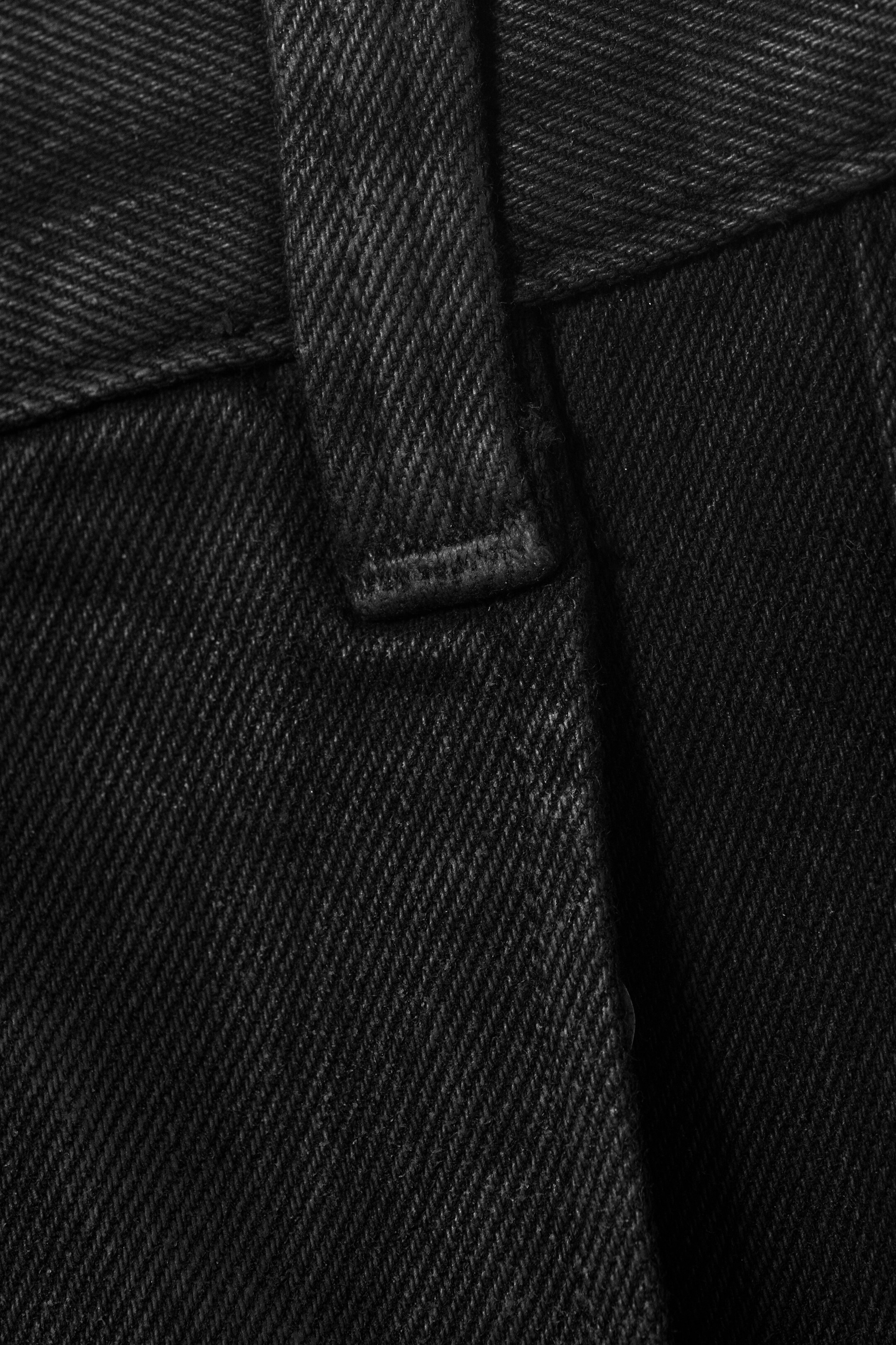 View larger image: A black garment is featuring a distinct twill weave texture. A belt loop, secured with matching black stitching, is visible at the top, and a vertical seam is running down the fabric, suggesting the front of the garment.