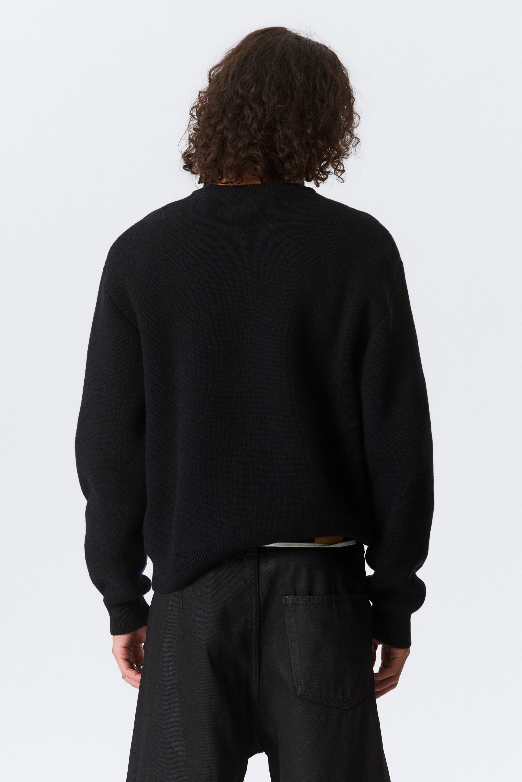 Se større billede: En sort strikket sweater bæres af modellen, med en ribbet halsudskæring, ribbet manchetter og en ribbet kant. Sweateren er lagdelt over et lyst farvet tøj, synligt ved kanten.