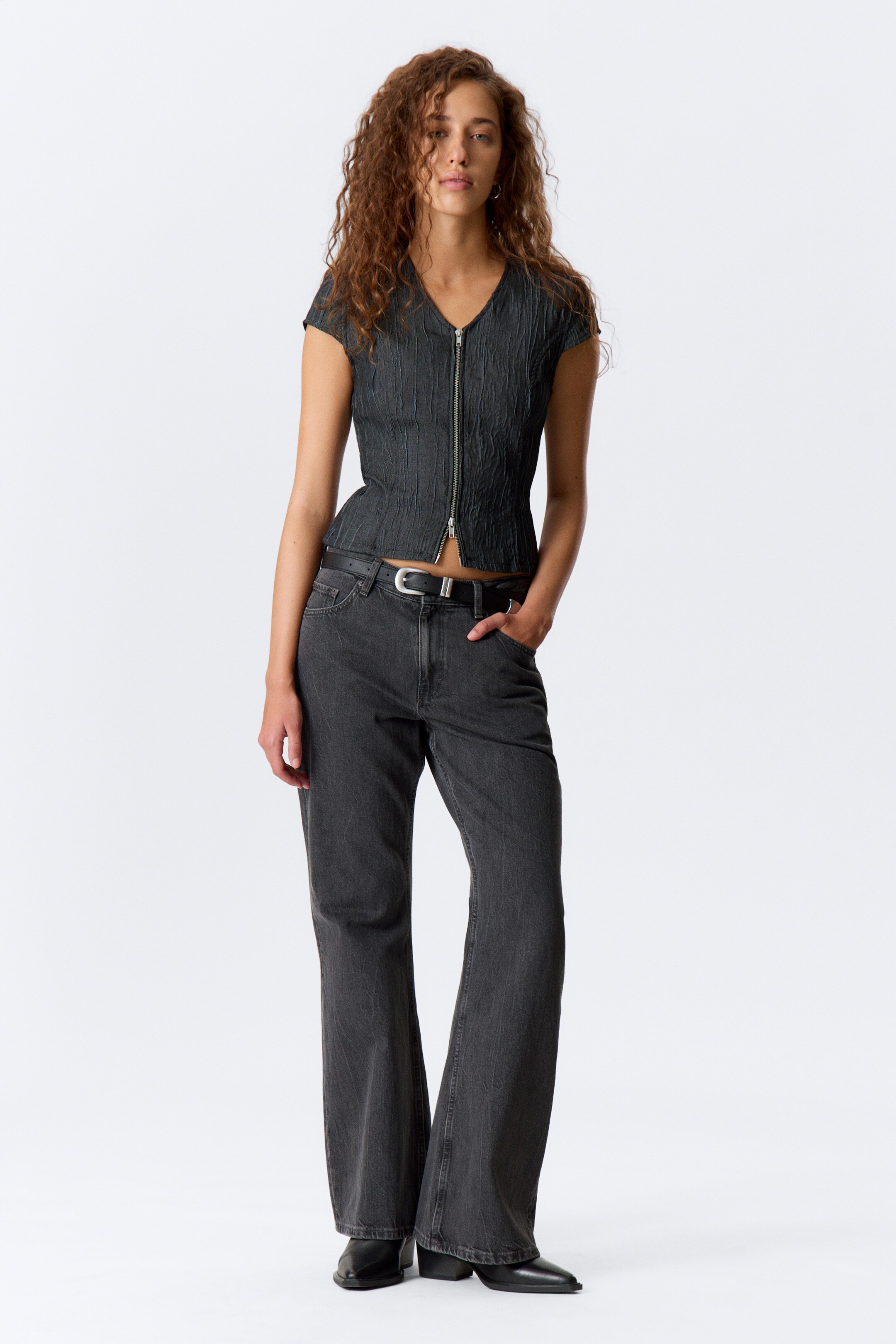 View larger image: A dark gray top is featuring a crinkled texture, a V-neck, short cap sleeves, and a full-length silver zipper down the front. It is being worn with high-waisted, relaxed-fit dark gray washed denim jeans, which are accessorized with a black belt and are flaring slightly at the hem.