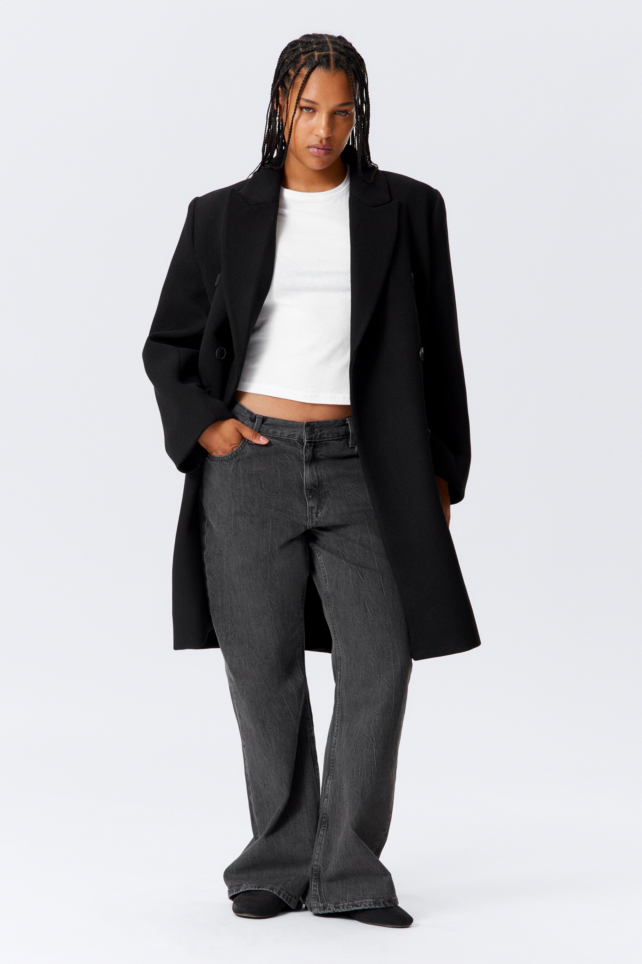 Ver imagem maior: Um casaco preto de abotoamento duplo está a ser apresentado num corte aberto e descontraído, com lapelas largas e estendendo-se até meio da coxa. Está sobreposto a um top branco cropped de gola redonda e jeans cinza escuro de perna larga, que apresentam um acabamento lavado e texturizado.