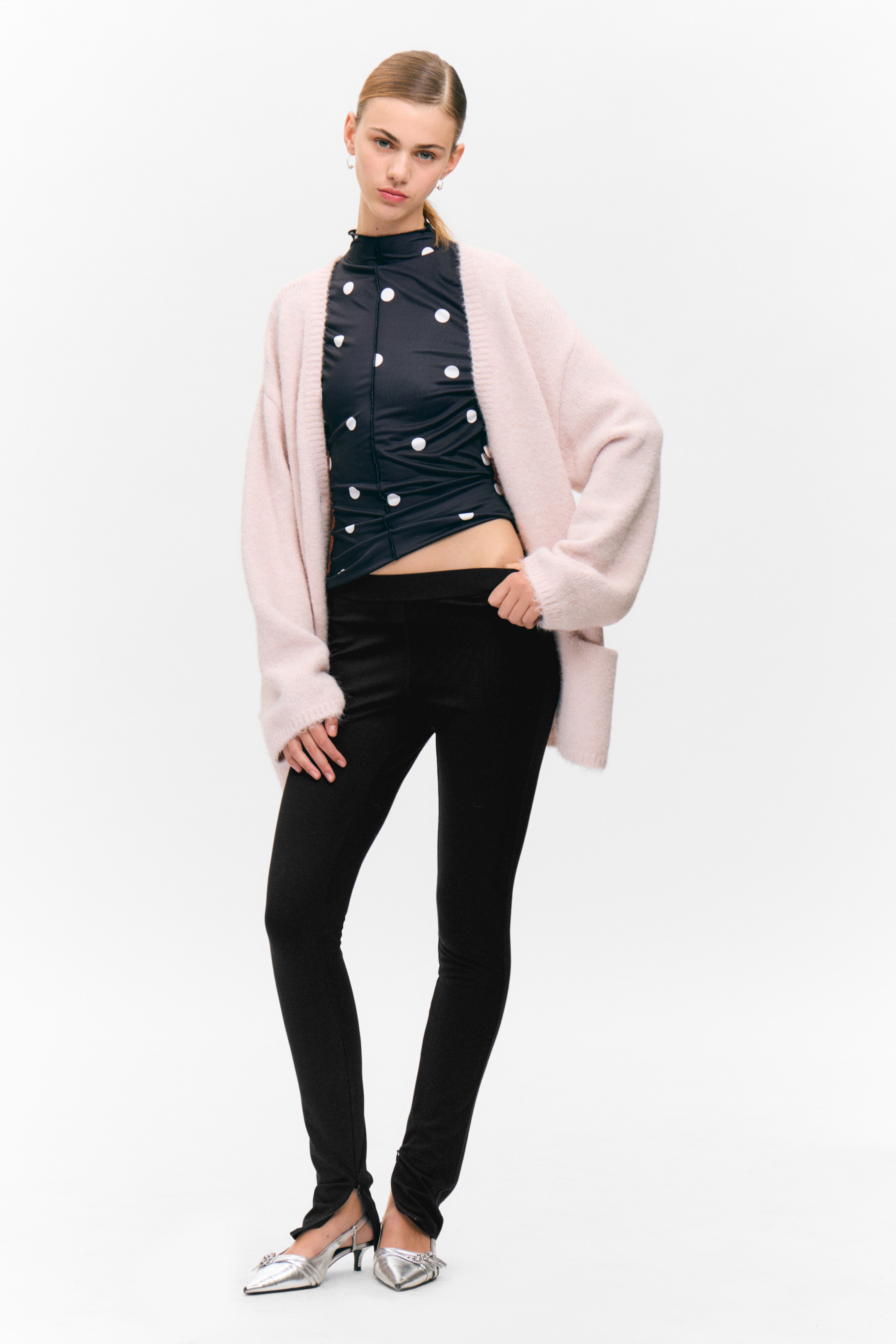 Visa större bild: Outfiten har en ljusrosa, öppen kofta med en mjuk, luddig textur som bärs över en svart, figurnära långärmad topp med vita prickar, med en rynkad framsida och en rynkad falsk polokrage. Svarta, smala byxor med en platt linning och subtila främre slitsar fullbordar looken.