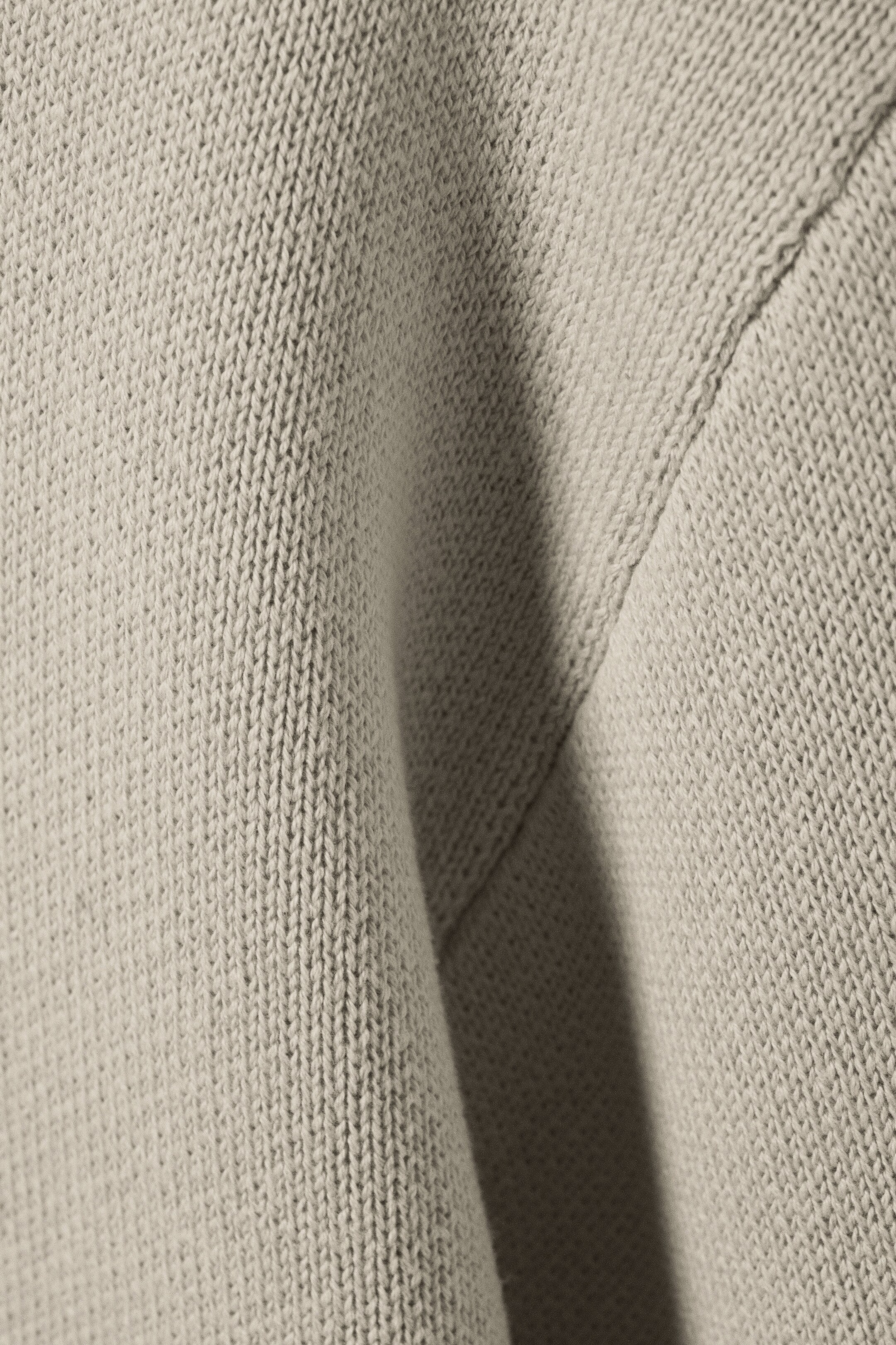 Größeres Bild anzeigen: Ein hellbeiger Strickstoff zeigt sein Jersey-Muster in einer Nahaufnahme, mit sichtbaren Falten, die Textur erzeugen, und einer Naht, die auf die Konstruktion des Kleidungsstücks hinweist.