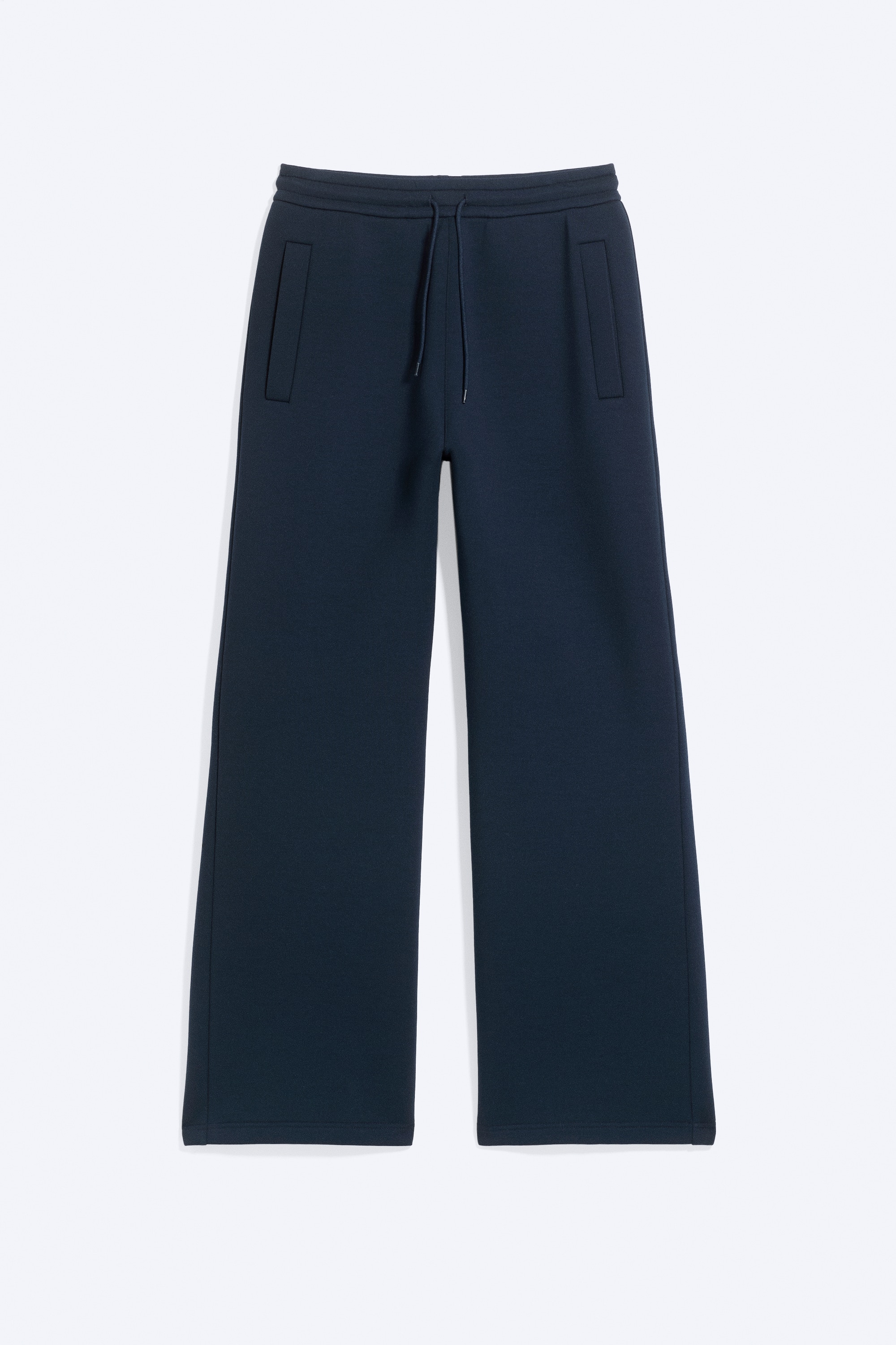 A pair of dark blue, loose-fit sweatpants in a structured scuba fabric is worn by the model, who is standing with hands resting in the side pockets. The sweatpants feature a relaxed silhouette and an elasticated drawstring waistband, and are styled with an olive green T-shirt, an open grey cardigan, and grey trainers.