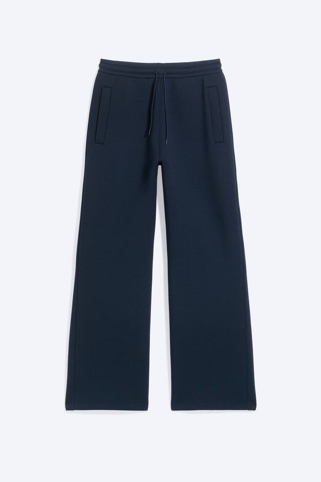 Ein Paar dunkelblaue, locker geschnittene Jogginghosen ist in Vorderansicht zu sehen, mit einem elastischen Bund mit Kordelzug und vertikalen Paspeltaschen. Die Hose fÃ¤llt in eine entspannte, weit geschnittene Silhouette und ist aus strukturiertem Scuba-Gewebe gefertigt. - 1