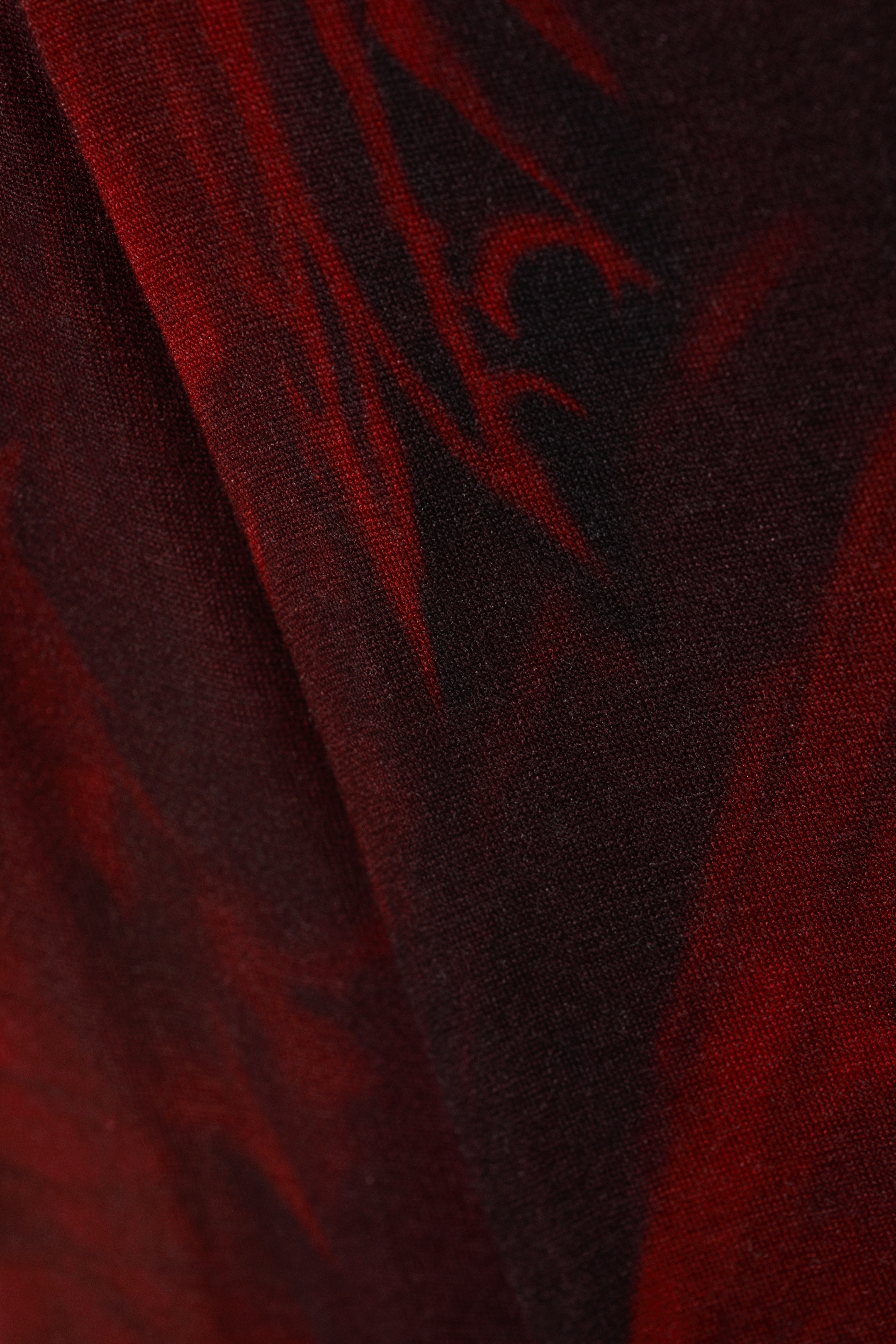 Größeres Bild anzeigen: Ein Textil zeigt ein abstraktes Muster aus tiefroten Formen auf einem schwarzen Hintergrund. Der Stoff zeigt subtile Falten und eine feine, strukturierte Webart.