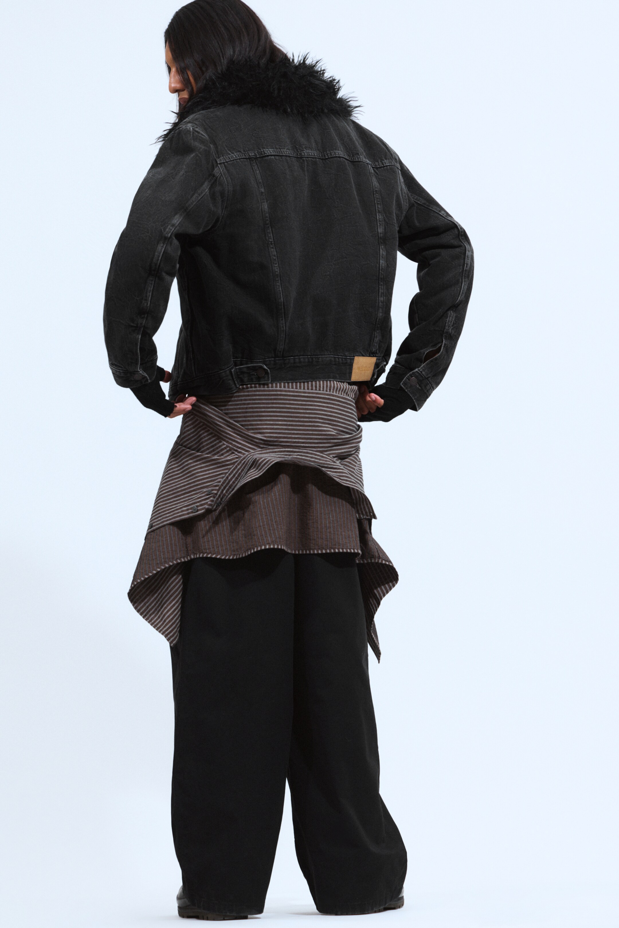 View larger image: A dark, washed denim jacket, featuring a black faux fur collar and a brown brand patch on the back hem, is being presented from a back view, styled over a striped brown and light-colored fabric garment that is being tied around the waist, creating an asymmetric, layered effect over black wide-leg pants. Black fingerless gloves are being worn on the hands.