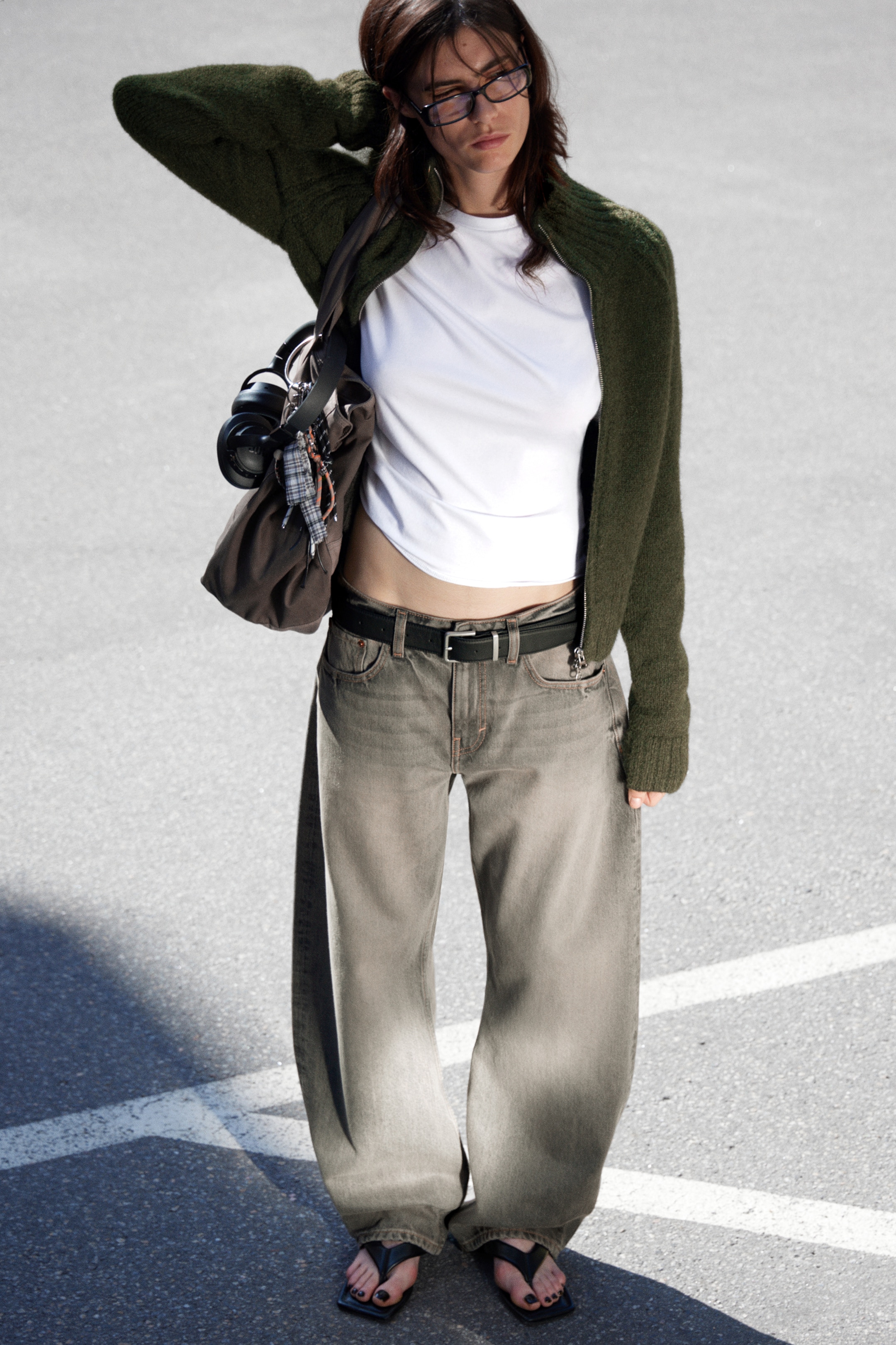 Grösseres Bild ansehen: Eine Person trägt eine dunkelgrüne, strukturierte Strickjacke, die offen ist, um ein weißes, kurzärmeliges Crop-Top darunter zu zeigen. Sie tragen auch ausgewaschene graue, weit geschnittene Jeans, die mit einem schwarzen Gürtel gesichert sind, und schwarze, quadratisch geformte Zehensandalen.