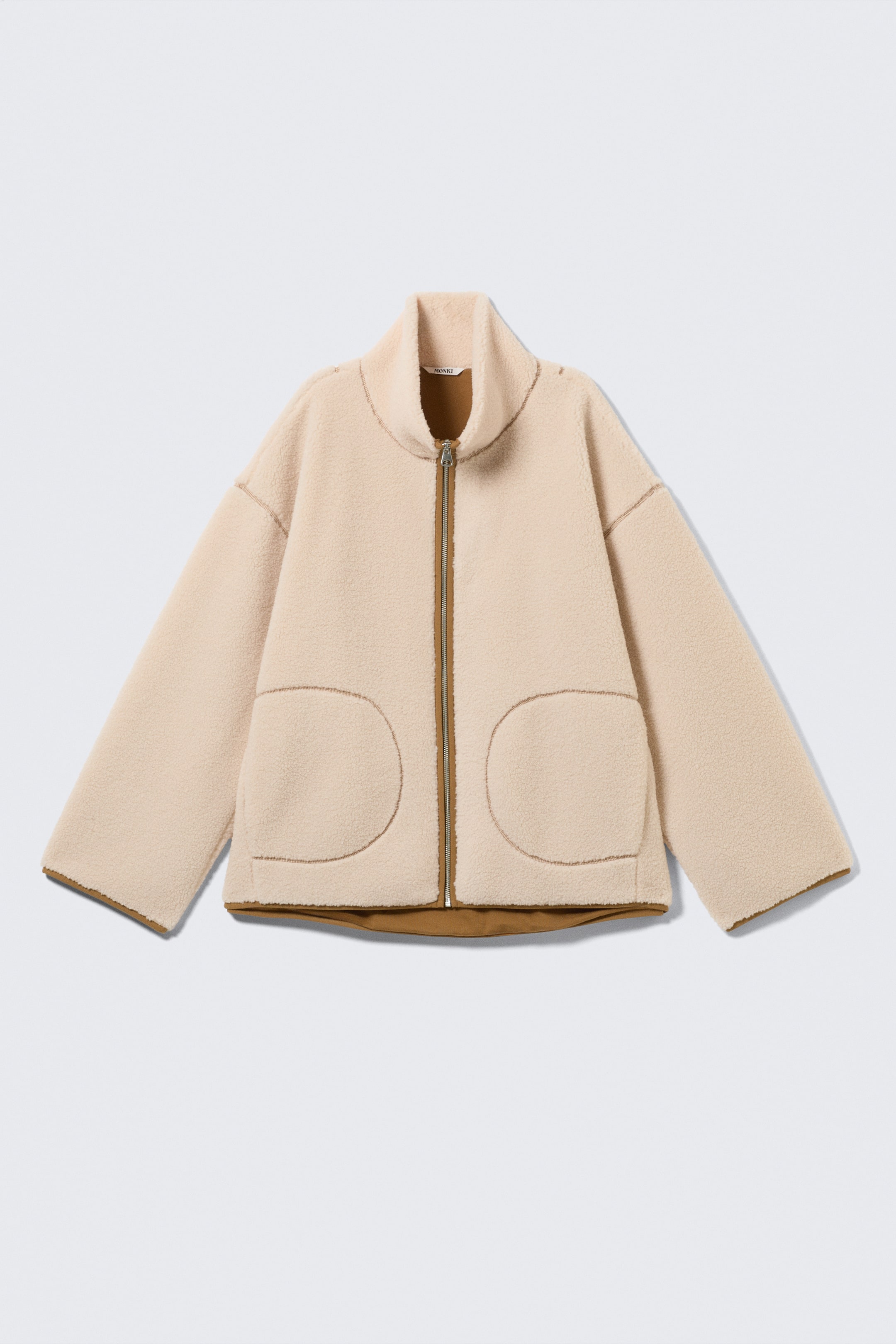 View larger image: FRONT view of a light beige pile jacket, showcasing a textured fleece material. It features a high collar, dropped shoulders, and two large, rounded patch pockets on the front. Brown piping outlines the full-length front zip, the pocket edges, the cuffs, and the hem, where a brown lining is also visible.