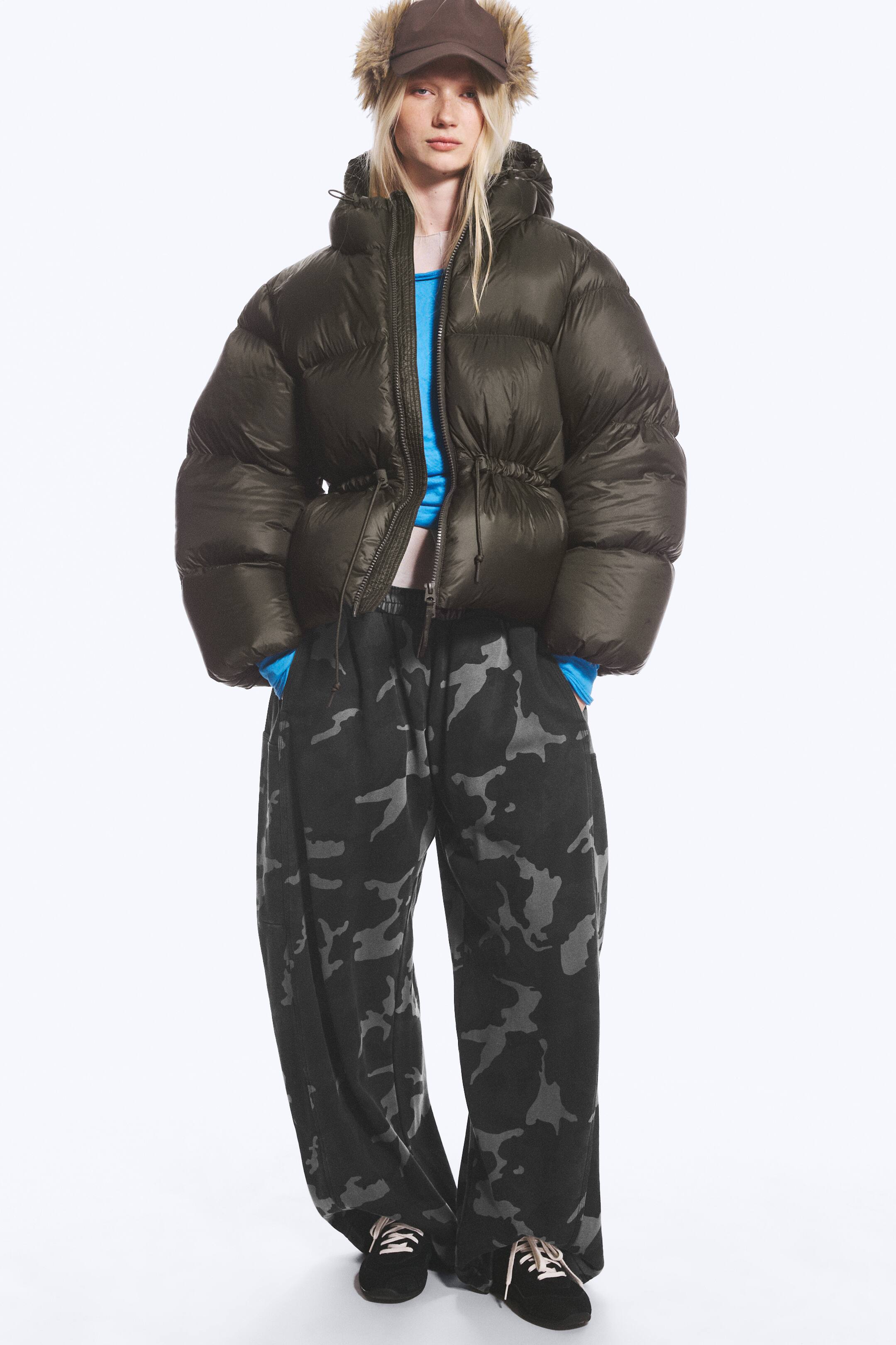 View larger image: An oversized, dark olive-green puffer jacket is being worn by the model, showcasing a voluminous quilted design with a partially unzipped front and a cinched drawstring waist. The jacket is styled with a bright blue long-sleeved top and dark, wide-leg camouflage trousers.