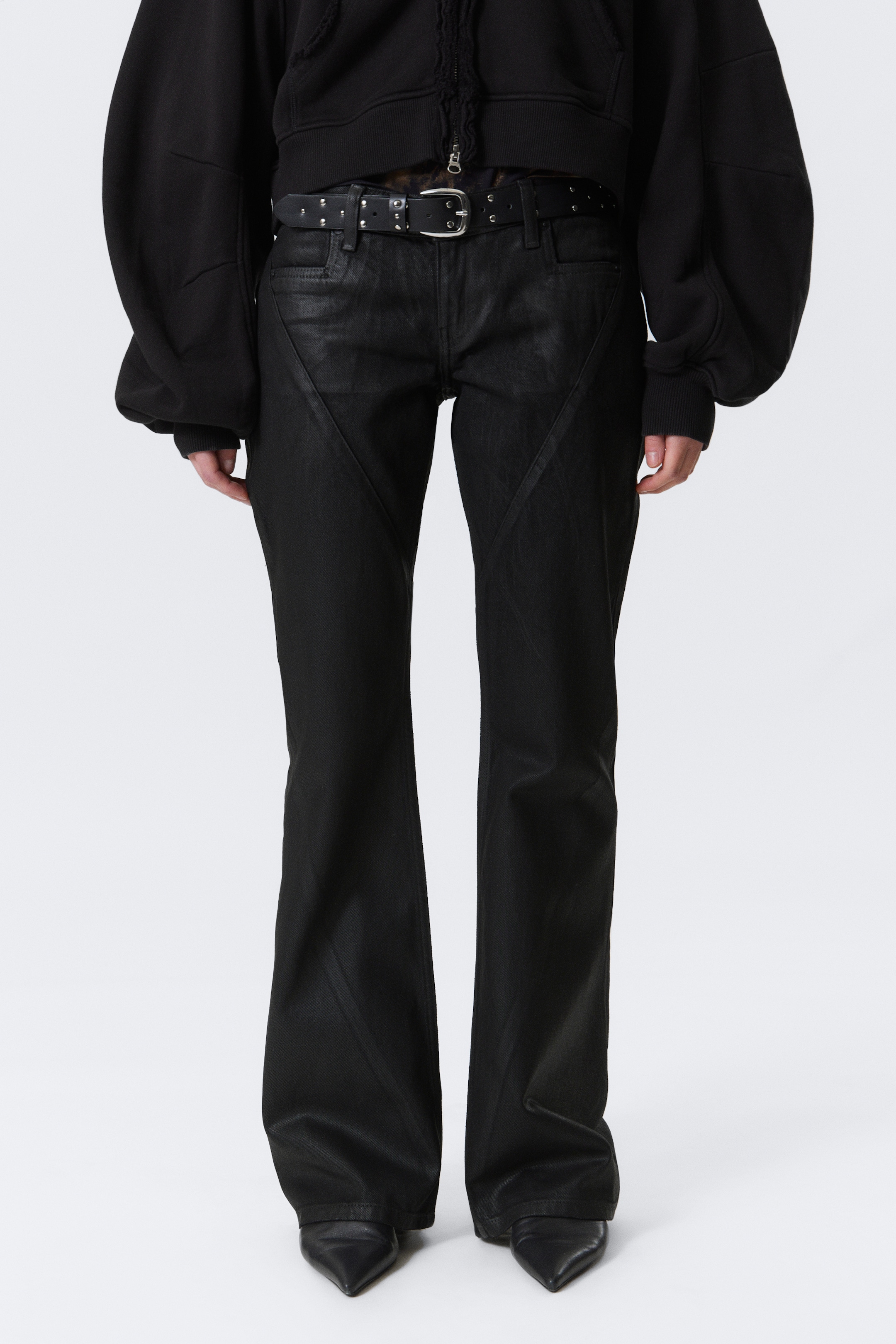 Größeres Bild ansehen: Schwarze ausgestellte Hosen werden von vorne präsentiert, mit einem subtilen Glanz und markanten V-förmigen Nähten auf der oberen Vorderseite. Die Hosen werden mit einem schwarzen, mit Nieten besetzten Gürtel und schwarzen spitzen Schuhen getragen.