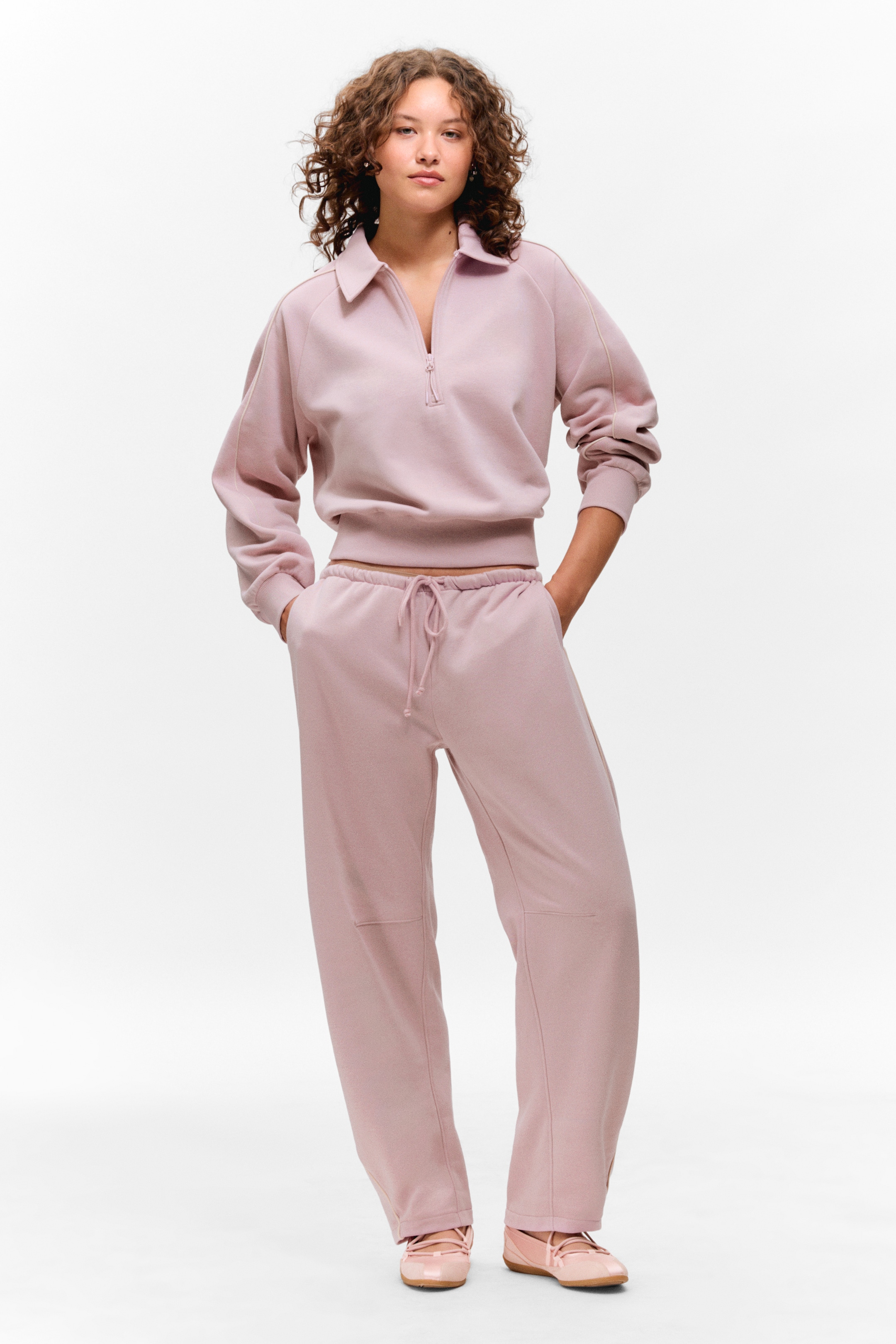 Vis større bilde: Et lyserosa loungewear-sett blir presentert, med modellen vendt fremover. Settet består av en halvglidelås genser med krage, raglanermer og ribbestrikkede mansjetter, og matchende avslappede joggebukser med snor i livet og synlige knepaneler.