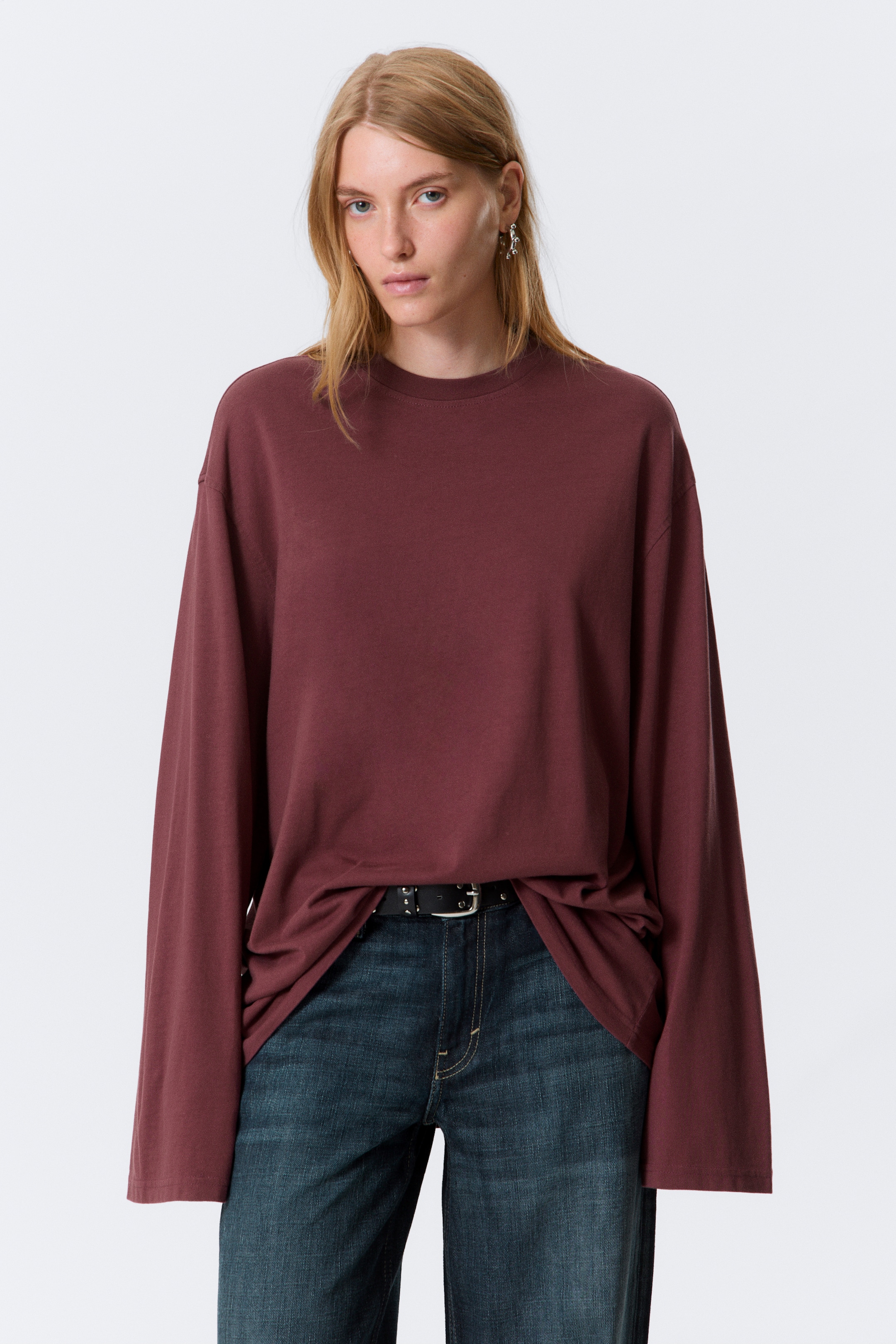 Visualizza immagine più grande: Un top bordeaux a maniche lunghe è indossato, caratterizzato da una vestibilità oversize e un girocollo. Le sue ampie maniche cadono morbide e l'orlo è parzialmente infilato nei jeans blu, mostrando il suo stile rilassato.