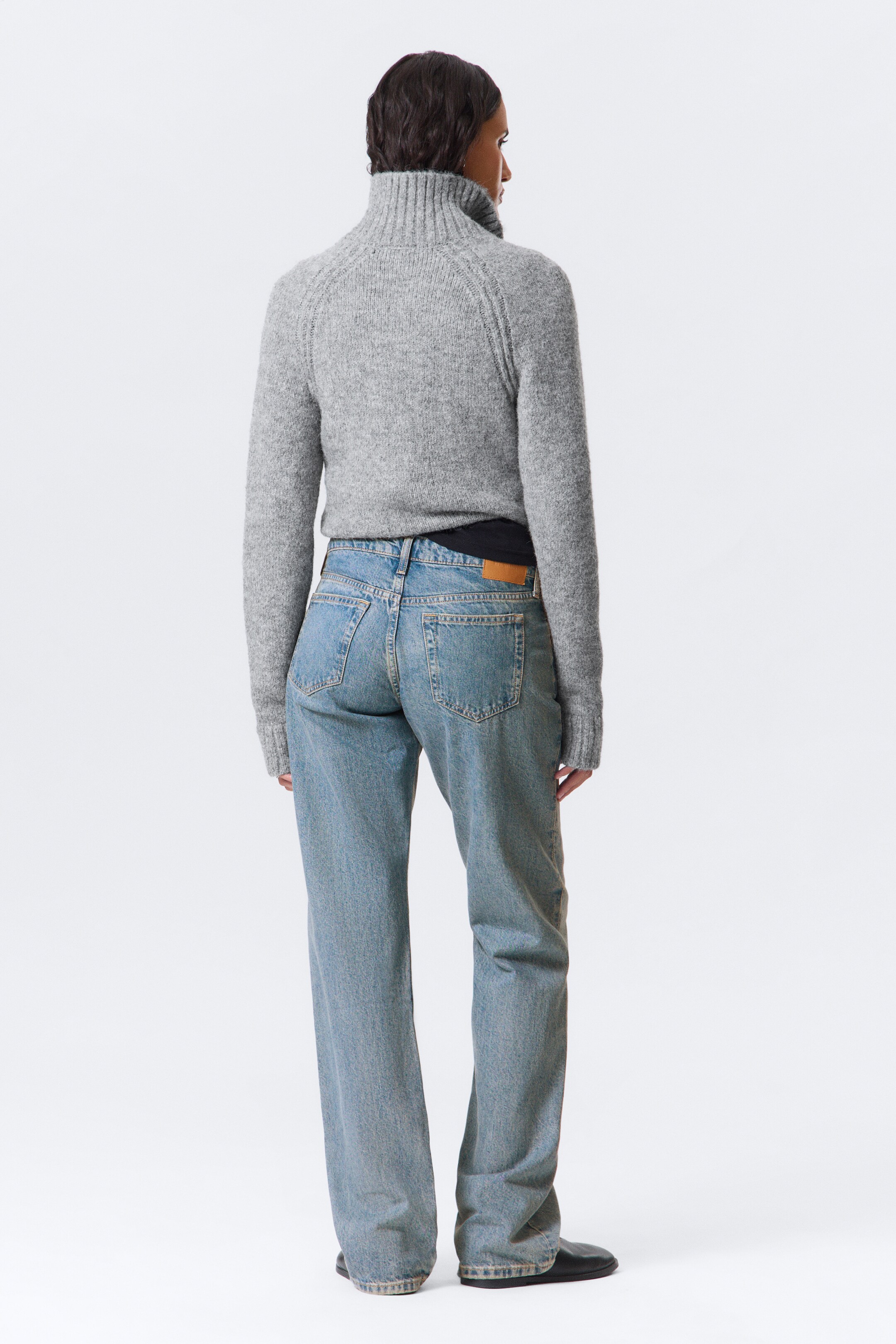 Agrandir l'image: Un pull gris tricoté avec un col montant côtelé, des manches raglan et des poignets côtelés est porté avec un jean bleu clair à coupe décontractée, vu de dos. Le jean présente des poches plaquées à l'arrière et une étiquette en cuir marron à la taille, avec une couche noire visible sous le pull.