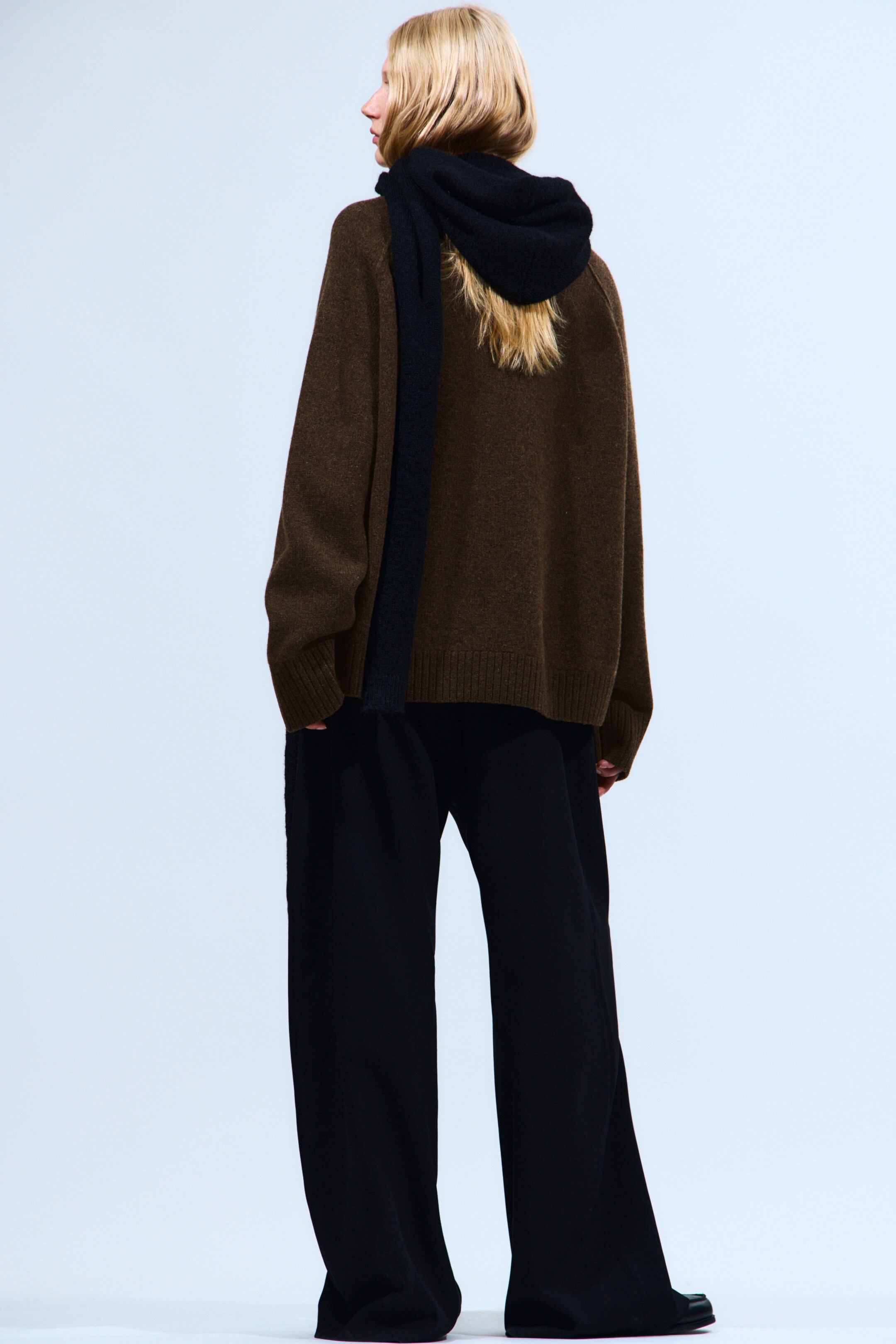 Agrandir l'image: Un pull en tricot marron surdimensionné, avec un ourlet côtelé, est porté, associé à un pantalon noir à jambes larges, le tout vu sous un angle arrière-droit. Une écharpe noire douce est drapée autour du cou et des épaules du pull, avec une extrémité tombant dans son dos.