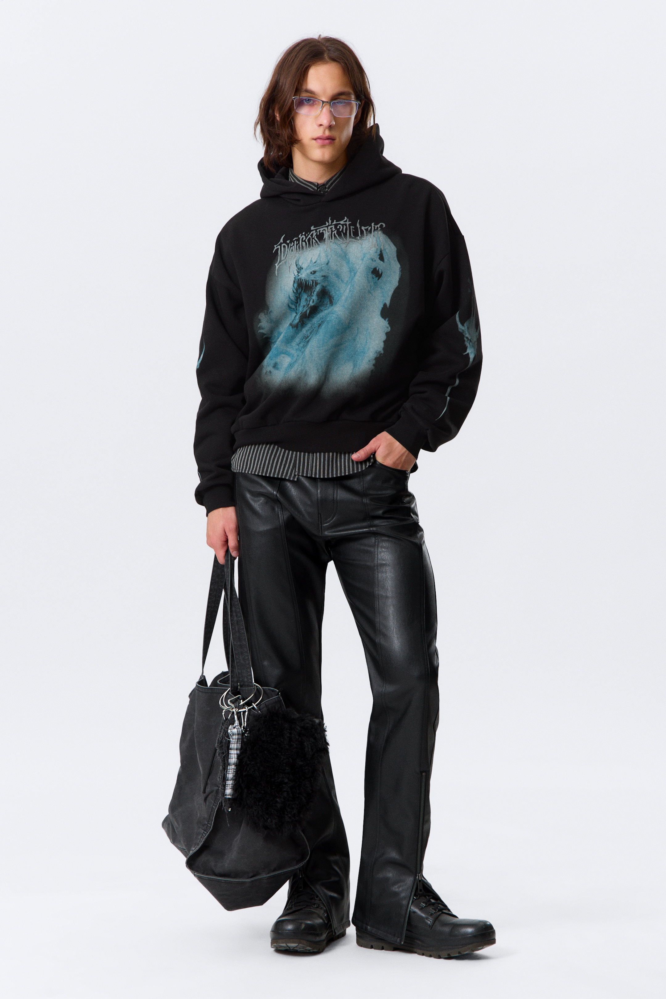 Agrandir l'image: Une tenue complète est présentée, mettant en vedette un sweat à capuche noir oversize avec un grand graphique bleu clair d'une créature ressemblant à un loup qui gronde sur la poitrine et des graphiques subtils sur les manches, superposé à une chemise à manches longues rayée. Un pantalon en faux cuir noir, stylisé avec des détails de coutures verticales et des fermetures éclair aux chevilles, complète le look avec des bottes de combat noires à lacets. Un sac noir à structure souple, accentué d'un porte-clés en fourrure noire et d'un autre à carreaux, est tenu en main.