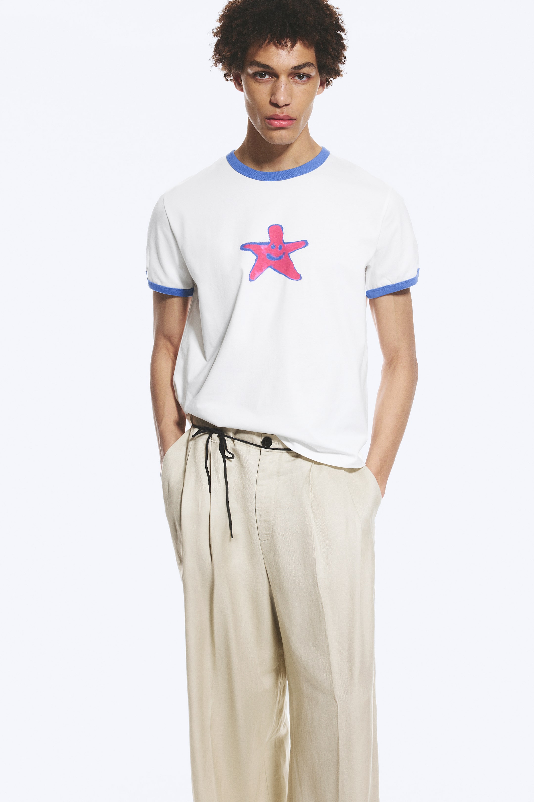 Agrandir l'image: Un t-shirt blanc à col ringer avec un col et des poignets côtelés bleus, orné d'un graphique d'étoile de mer rose et bleue sur la poitrine. Il est porté avec un pantalon plissé beige clair doté d'une taille à cordon de serrage noir.