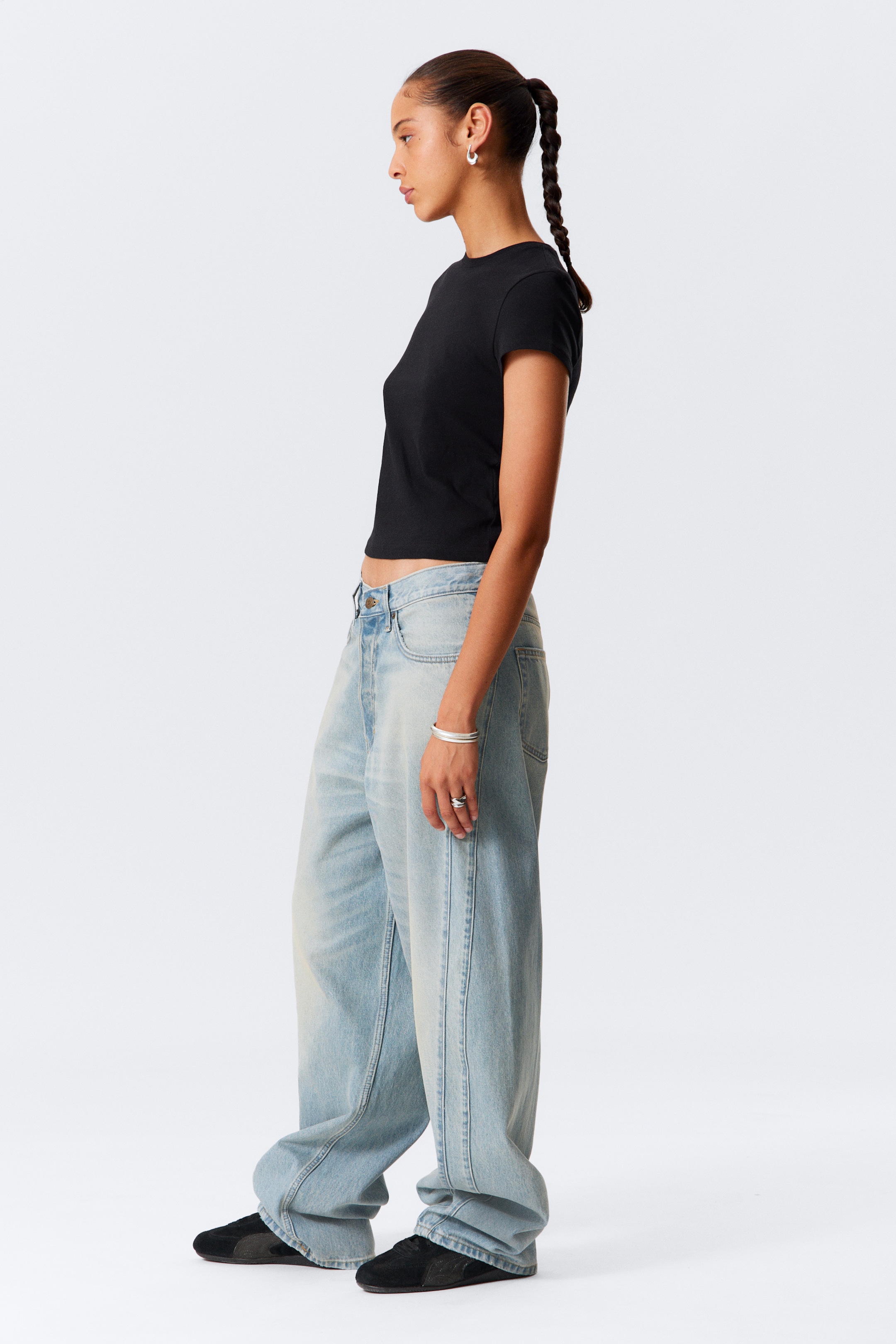 Visa större bild: En svart, figursydd, kortärmad t-shirt med rund hals bärs tillsammans med ljusa, löst sittande denimjeans. T-shirten är kort och jeansen har en pösig, vid design, båda sedda från sidan.