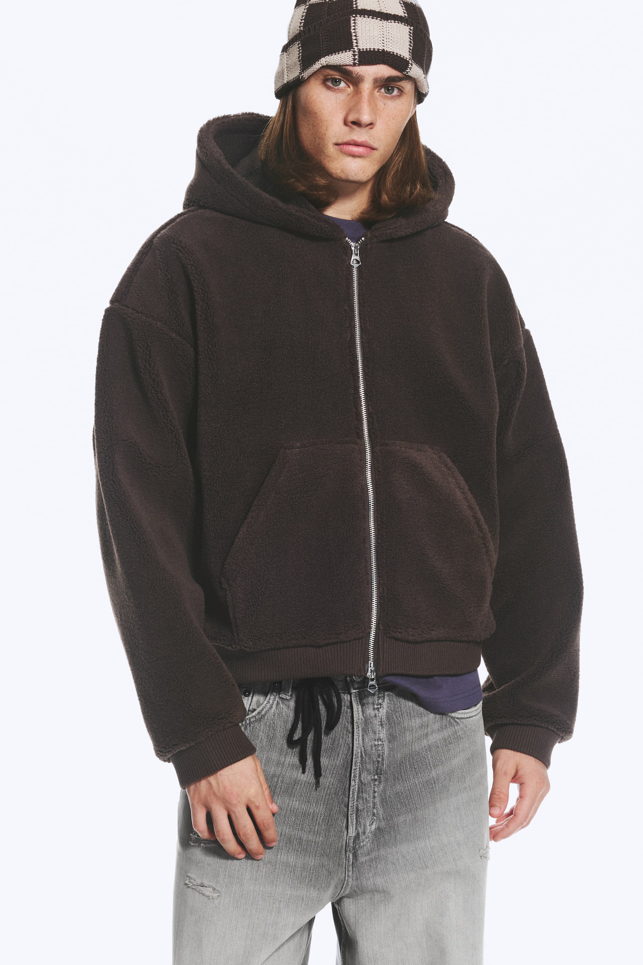 View larger image: A dark brown fleece hoodie featuring a silver two-way front zip, a hood, and ribbed cuffs is being worn by the model. The product is styled with grey jeans and a checked beanie.
